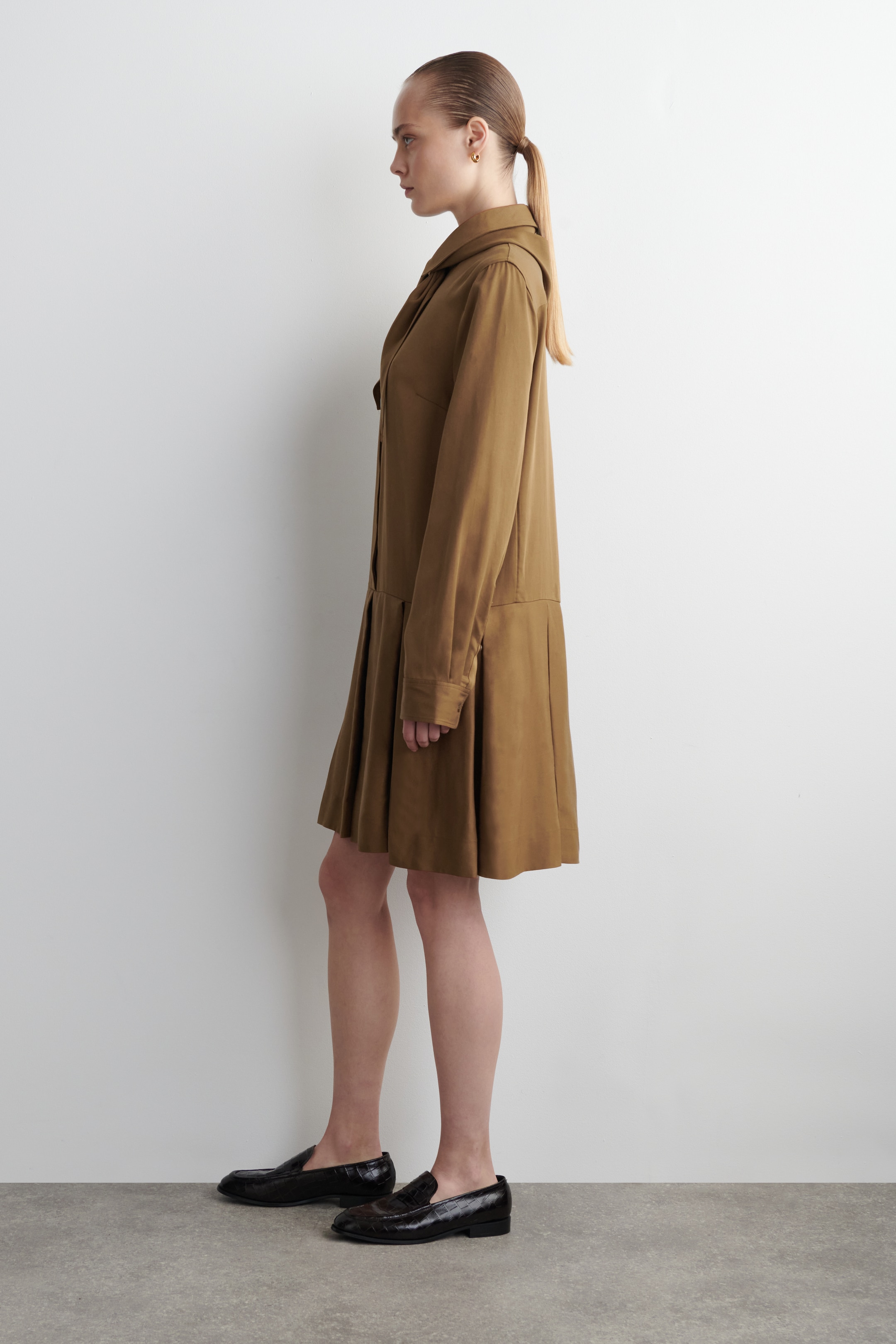 Ver imagem maior: O vestido mini ocre é mostrado a partir da esquerda, apresentando uma gola pontiaguda, mangas compridas e um elemento de lenço tonal drapeado em torno do pescoço. O vestido é desenhado com uma silhueta de cintura baixa e pregas profundas na saia.