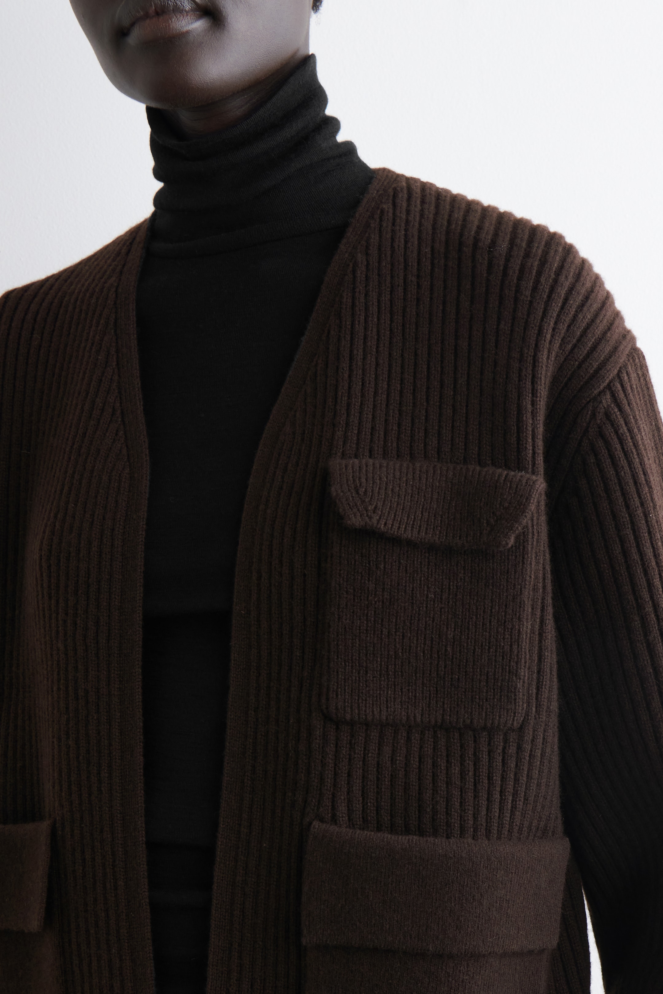 Grotere afbeelding bekijken: Een bruine geribbelde gebreide cardigan wordt van voren gepresenteerd, open gedragen over een zwarte coltrui. Het heeft opvallende verticale ribbels en twee zichtbare klepzakken, één op de borst en een andere lager op het lichaam.