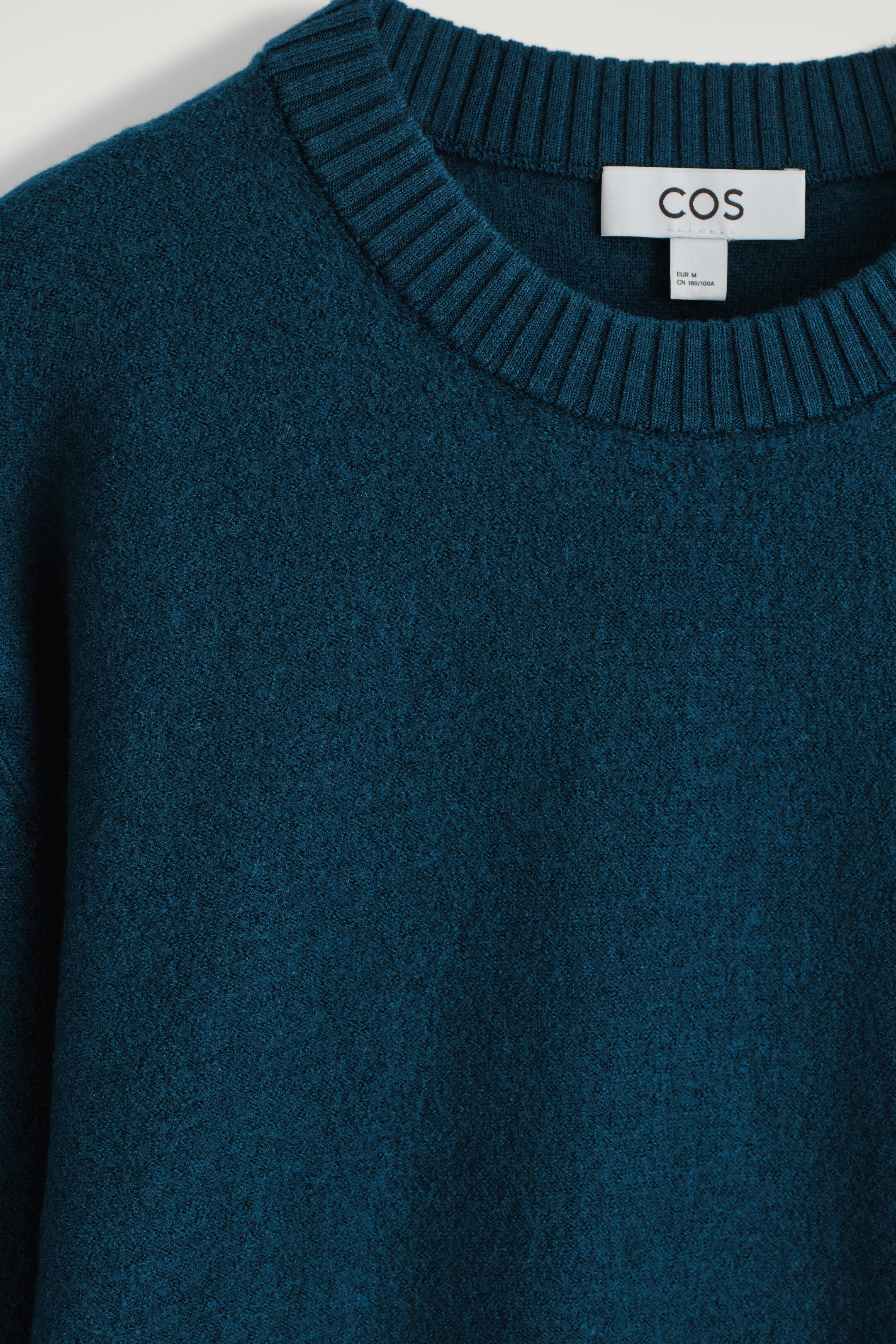 View larger image: A dark teal wool sweater is prominently displayed from the front, featuring a classic crew neck with a wide ribbed trim. Its knitted fabric is showing a soft, textured appearance.