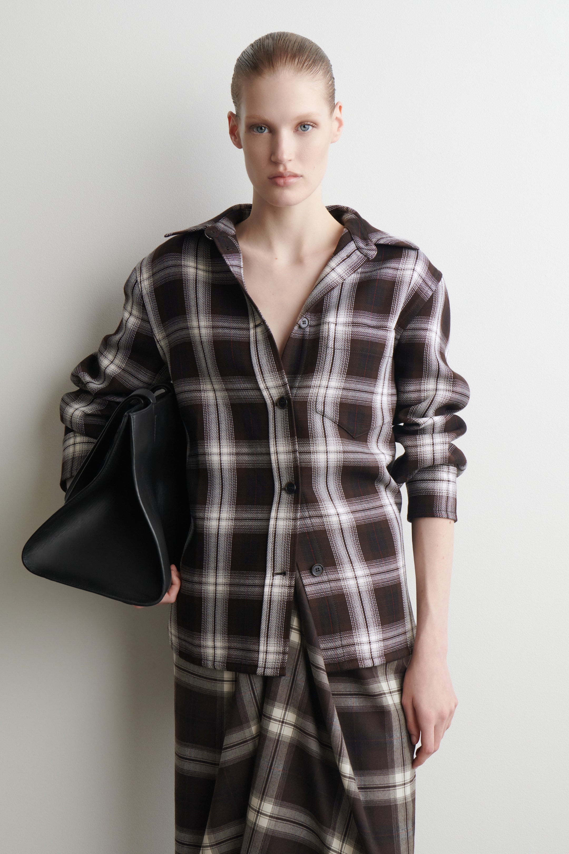 Agrandir l'image: Une chemise à carreaux marron, blanc cassé et foncé est portée, avec un col pointu, une poche poitrine unique et une coupe décontractée et ample, déboutonnée en haut. Elle est associée à une jupe à carreaux coordonnée, présentant un design drapé et asymétrique. Un sac structuré noir est tenu sous le bras gauche.