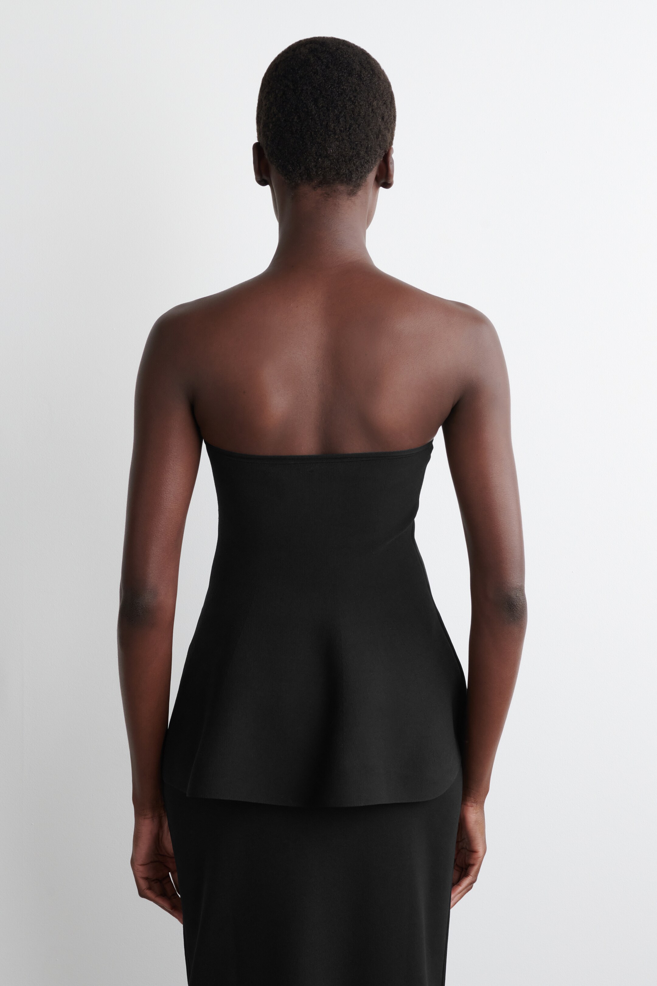 Ver imagen más grande: Un top negro sin tirantes con peplum se está viendo desde atrás. Presenta un corpiño ajustado y un dobladillo de peplum acampanado, y se superpone sobre una falda negra.