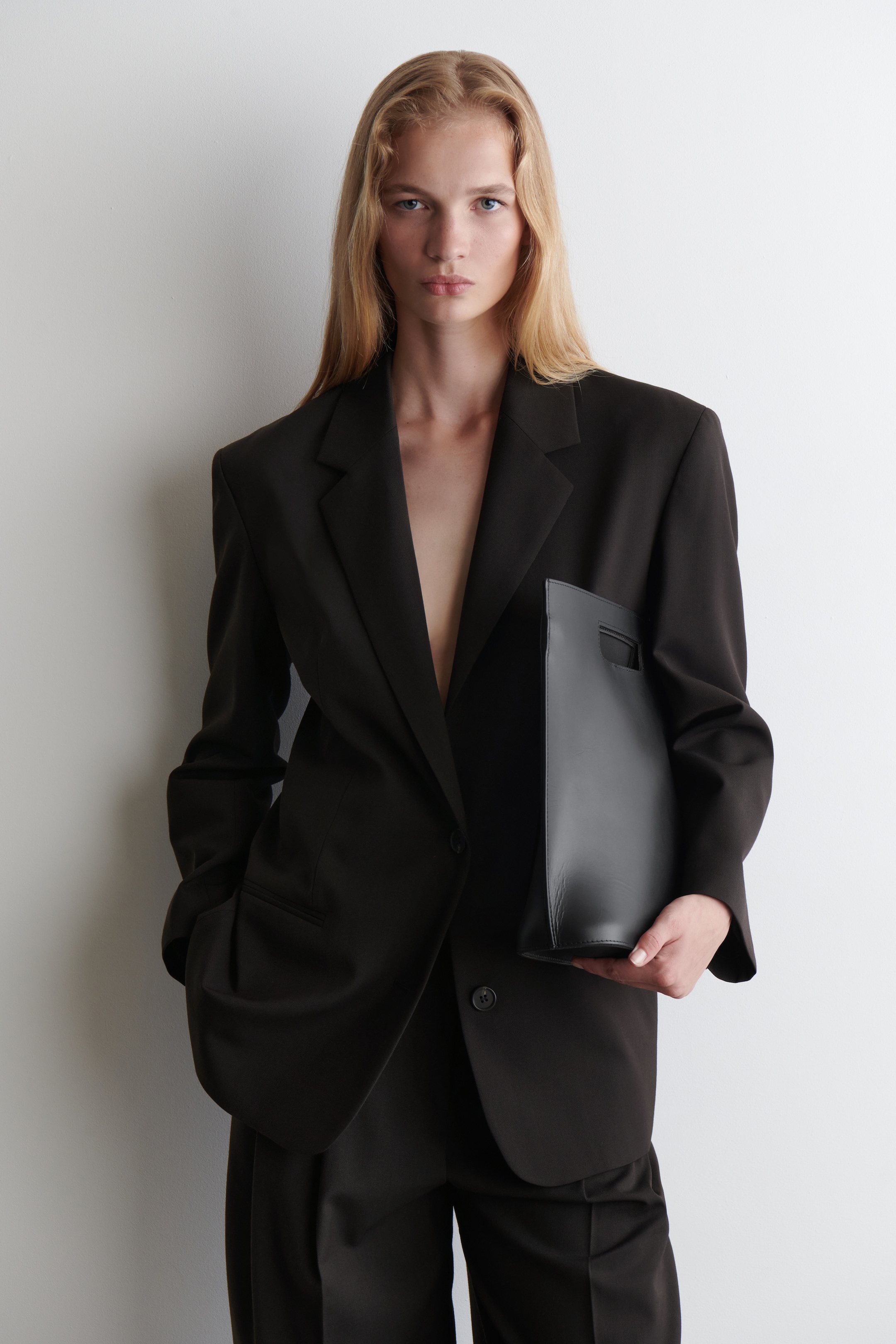 Agrandir l'image: Un blazer oversize de couleur sombre avec des revers crantés et un seul bouton visible est porté ouvert avec un pantalon large assorti, tandis qu'un sac rectangulaire noir avec une poignée découpée est tenu contre le blazer.