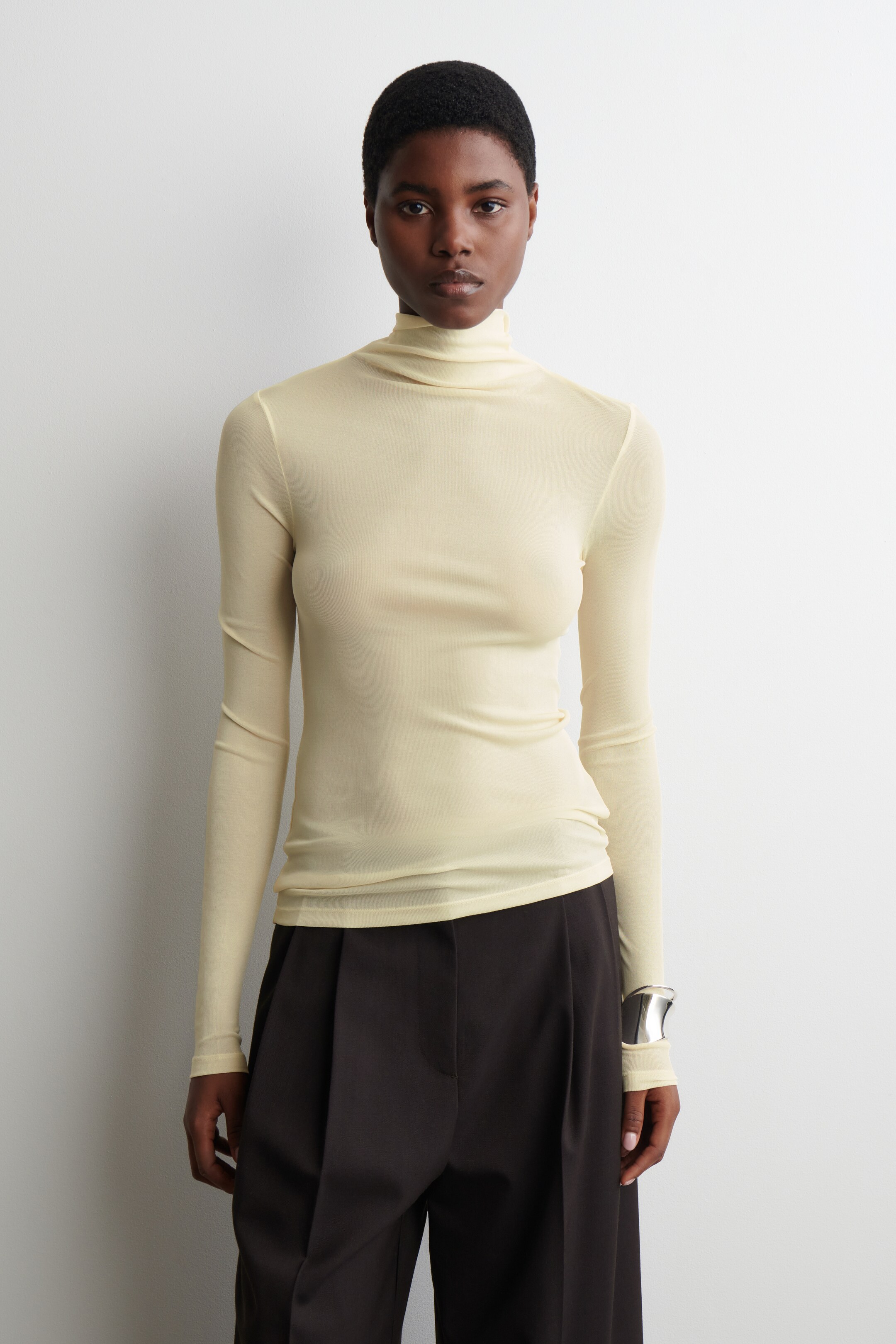 Visualizza immagine più grande: Una maglia giallo chiaro, semi-trasparente, aderente, con collo alto e maniche lunghe è indossata dalla modella. La maglia si estende sopra pantaloni scuri.