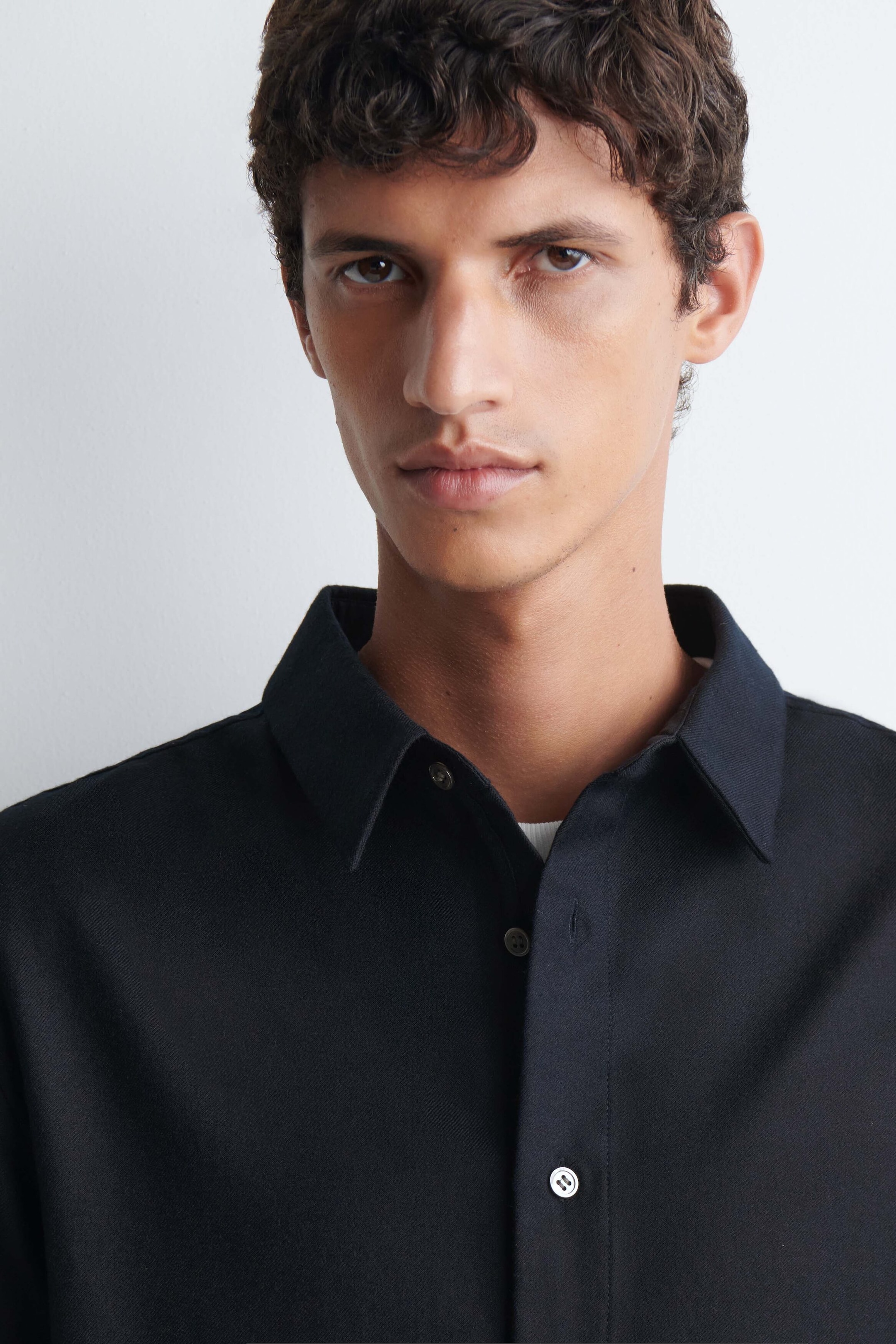 Das Model trägt ein schwarzes, leger geschnittenes Hemd aus Wollflanell mit klassischem spitzem Kragen und durchgehender Knopfleiste. Ein weißes Unterhemd ist am offenen Kragenansatz leicht sichtbar. Das Model trägt ein schwarzes, leger geschnittenes Hemd aus Wollflanell mit klassischem spitzem Kragen und durchgehender Knopfleiste. Ein weißes Unterhemd ist am offenen Kragenansatz leicht sichtbar.