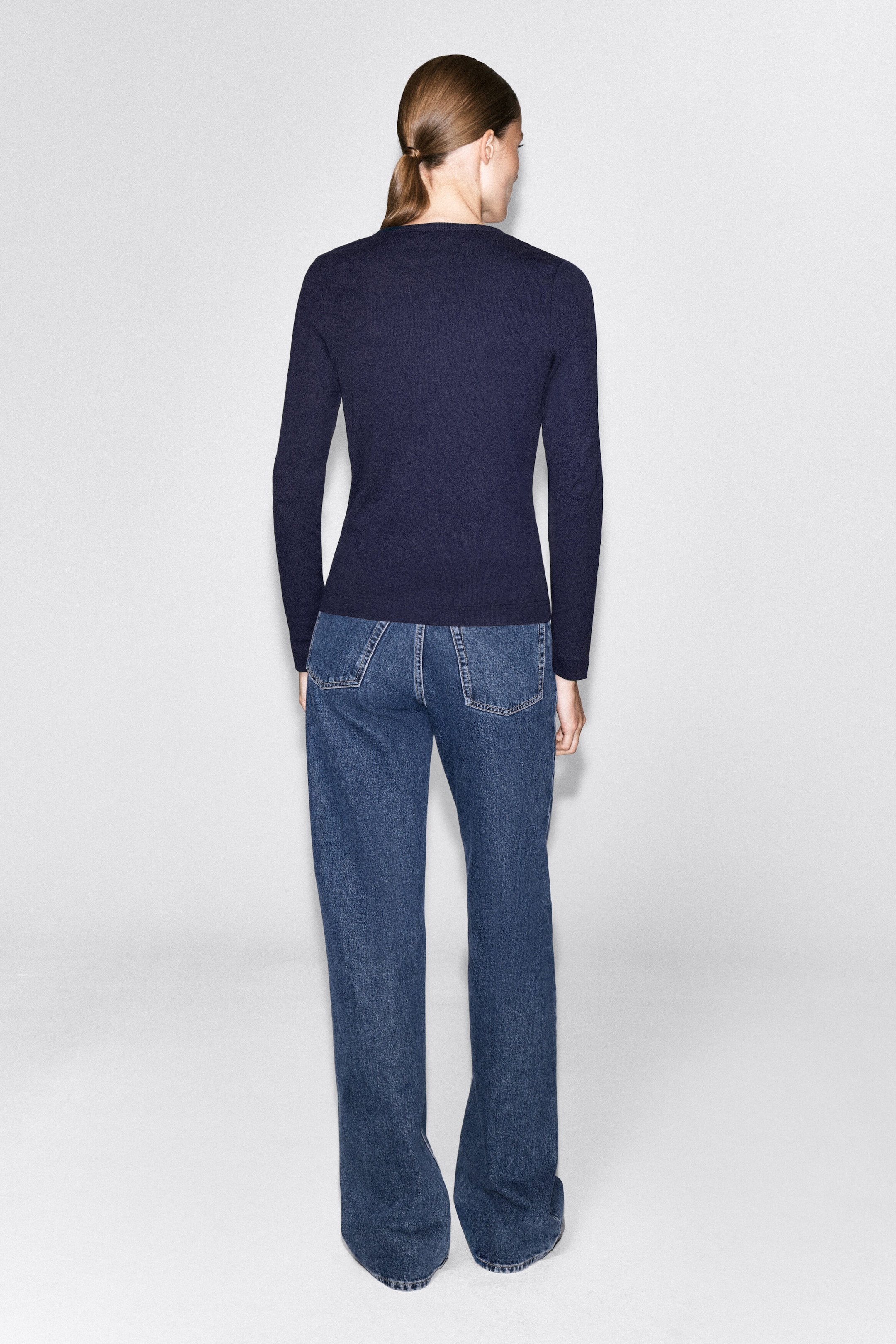 The model is wearing a navy blue ribbed cotton henley top, showing the back of its long-sleeved, slim-fit design. The top is styled with a pair of blue jeans. The model is wearing a navy blue ribbed cotton henley top, showing the back of its long-sleeved, slim-fit design. The top is styled with a pair of blue jeans.