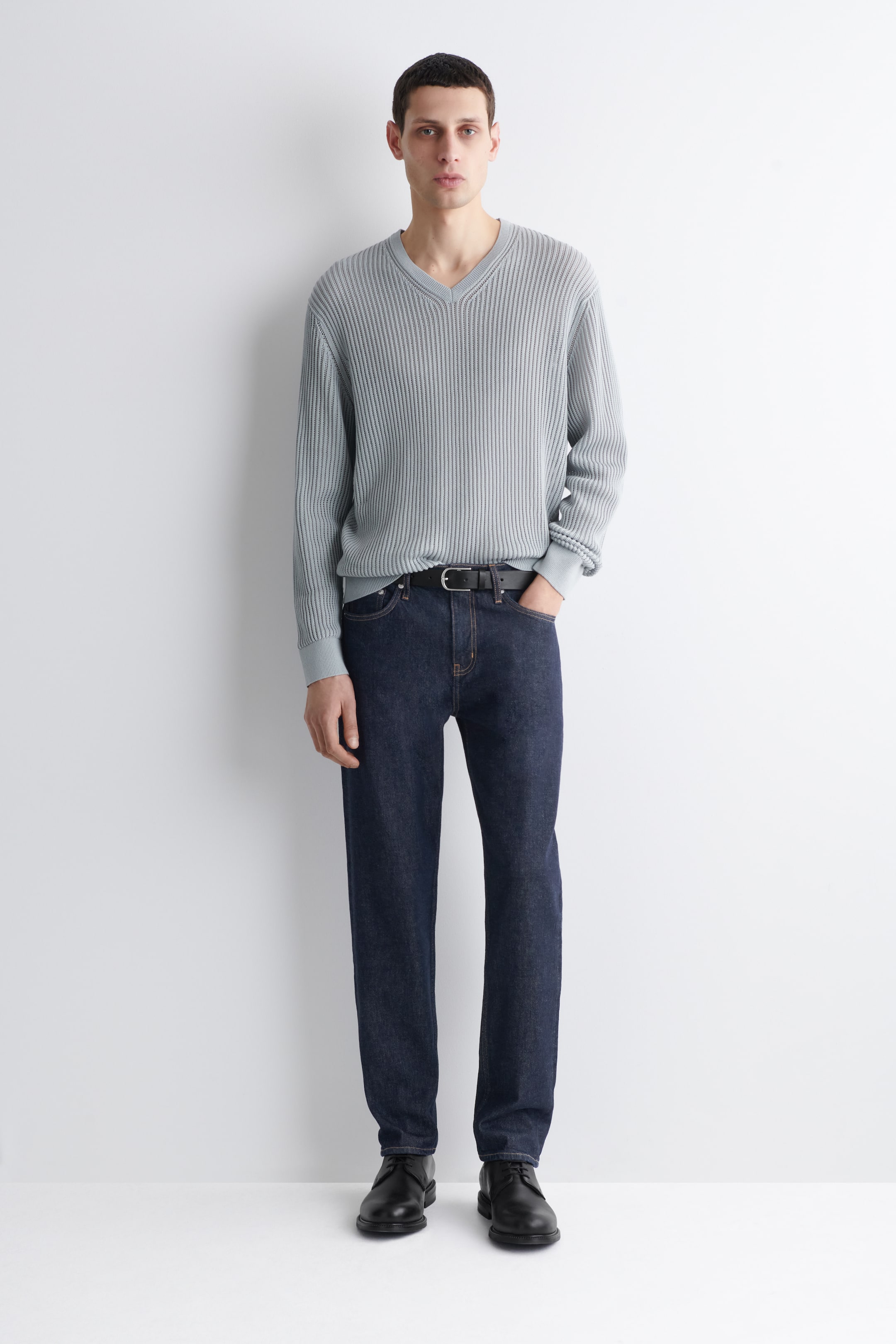 Visualizza immagine più grande: Un maglione grigio chiaro con scollo a V viene presentato frontalmente, caratterizzato da una lavorazione a maglia a coste su tutta la superficie, maniche lunghe con polsini a coste e un orlo a coste infilato nei jeans scuri.