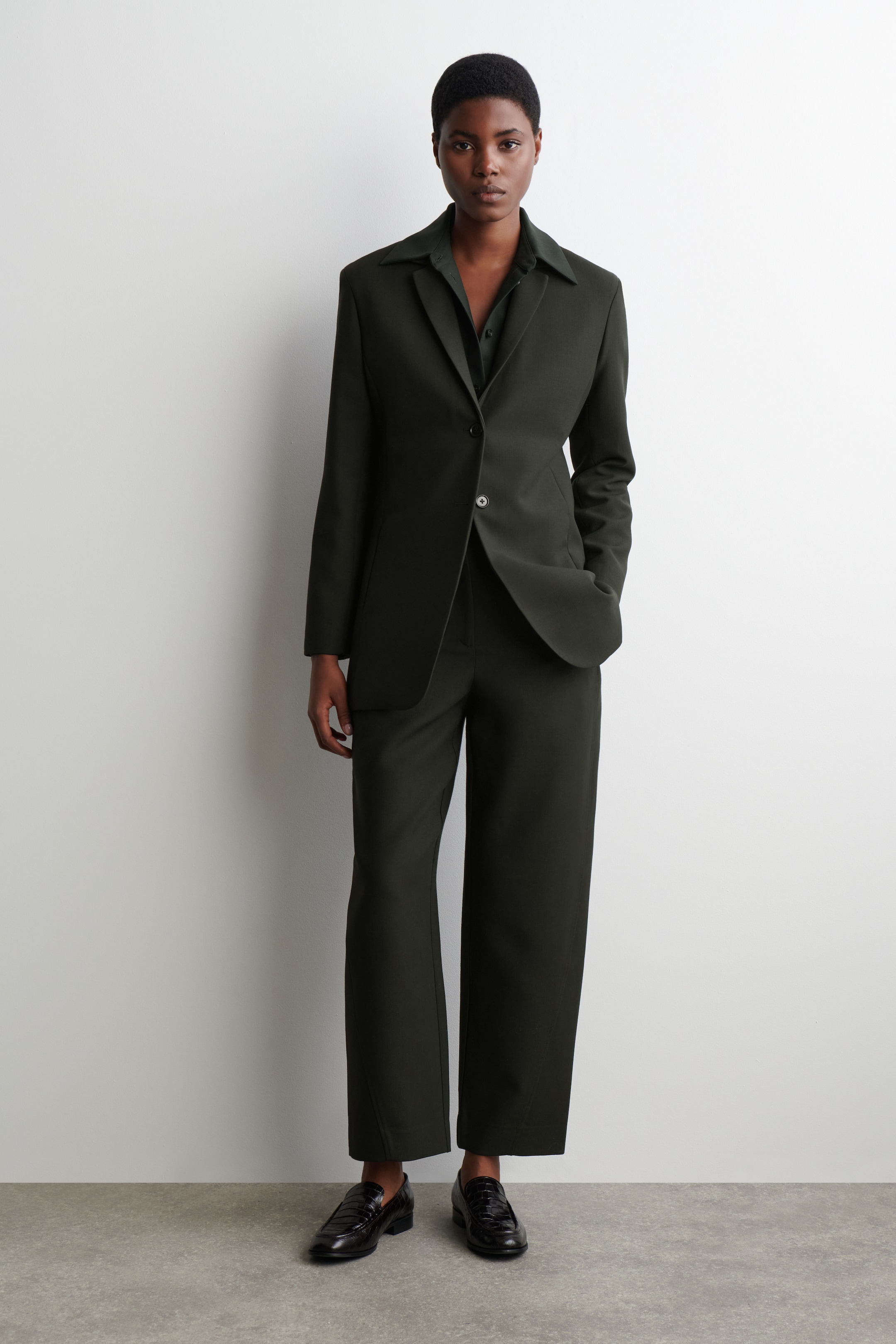 Agrandir l'image: Un costume vert foncé à simple boutonnage est présenté de face. Le blazer est doté de revers crantés, d'une fermeture à un seul bouton de couleur claire et d'épaules structurées, porté sur une chemise à col vert plus foncé. Un pantalon large assorti complète le blazer, associé à des mocassins texturés foncés.