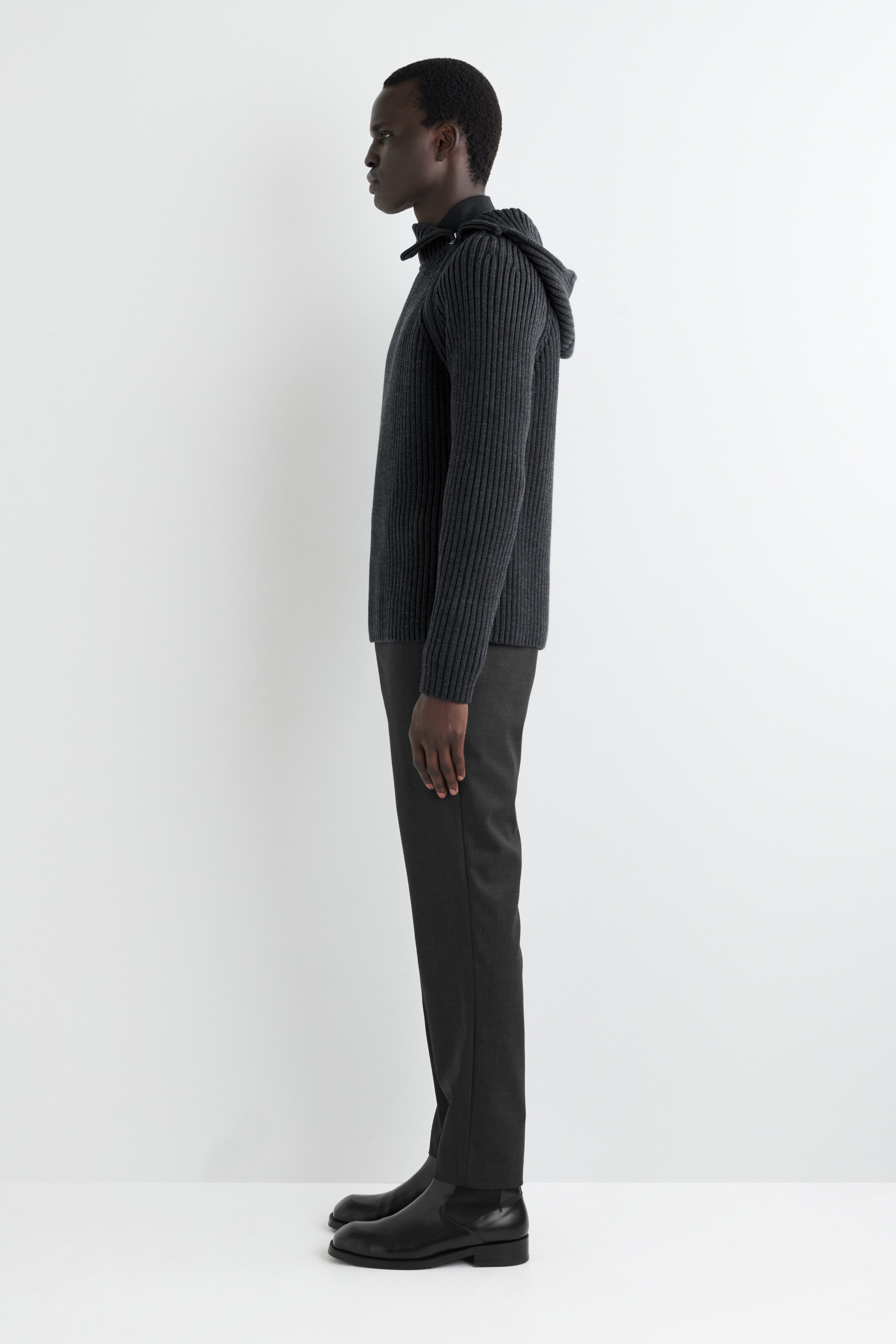 Agrandir l'image: Un pull à capuche gris foncé à cotes est porté, avec un col à cordon de serrage et une capuche visible à l'arrière, superposé à une chemise foncée. Il est assorti à un pantalon gris foncé et des bottines Chelsea noires, l'ensemble étant présenté de profil complet.