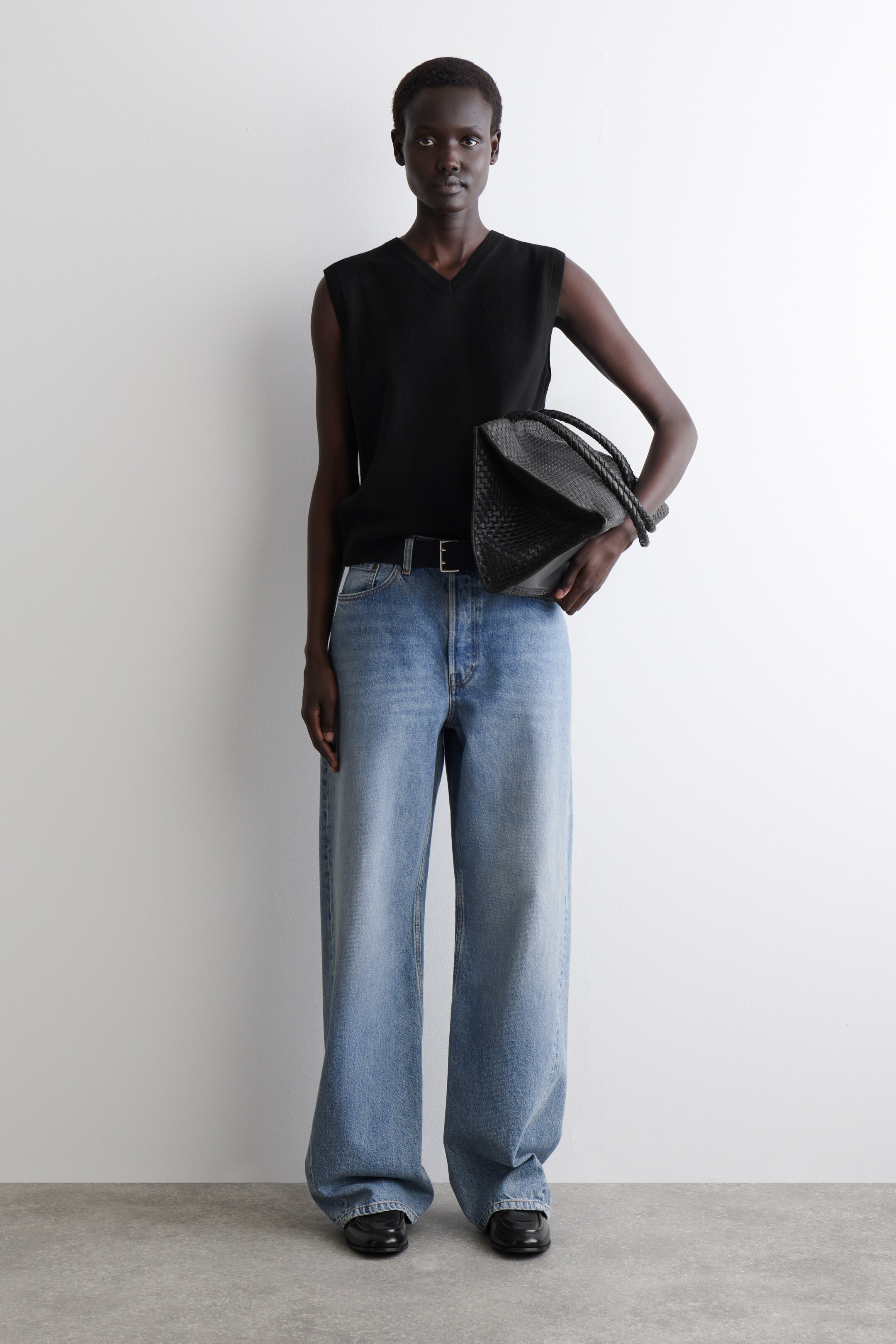 Visualizza immagine più grande: L'intero outfit viene presentato da un'angolazione frontale, con un top nero senza maniche a V in maglia che viene indossato infilato in jeans di denim a gamba larga e lavaggio chiaro. I jeans sono stretti con una cintura nera e vengono indossati mocassini neri piatti. Una borsa intrecciata nera viene tenuta al lato.