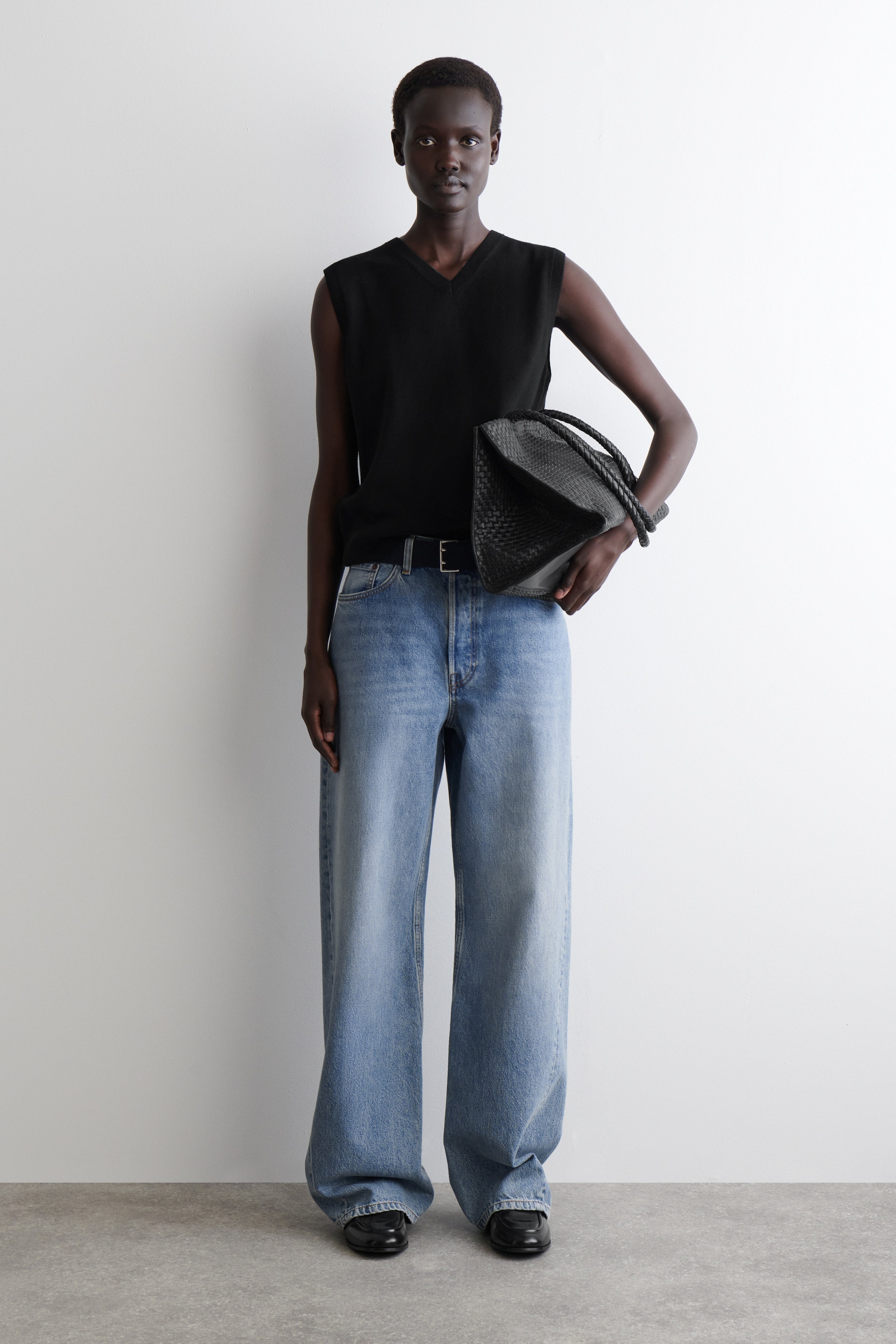 Größeres Bild ansehen: Das komplette Outfit wird aus einem frontalen Winkel präsentiert und zeigt ein schwarzes ärmelloses V-Ausschnitt-Stricktop, das in hell gewaschene, weit geschnittene Jeans gesteckt wird. Die Jeans werden mit einem schwarzen Gürtel geschnürt, und schwarze flache Loafer werden getragen. Eine schwarze gewebte Handtasche wird an der Seite gehalten.