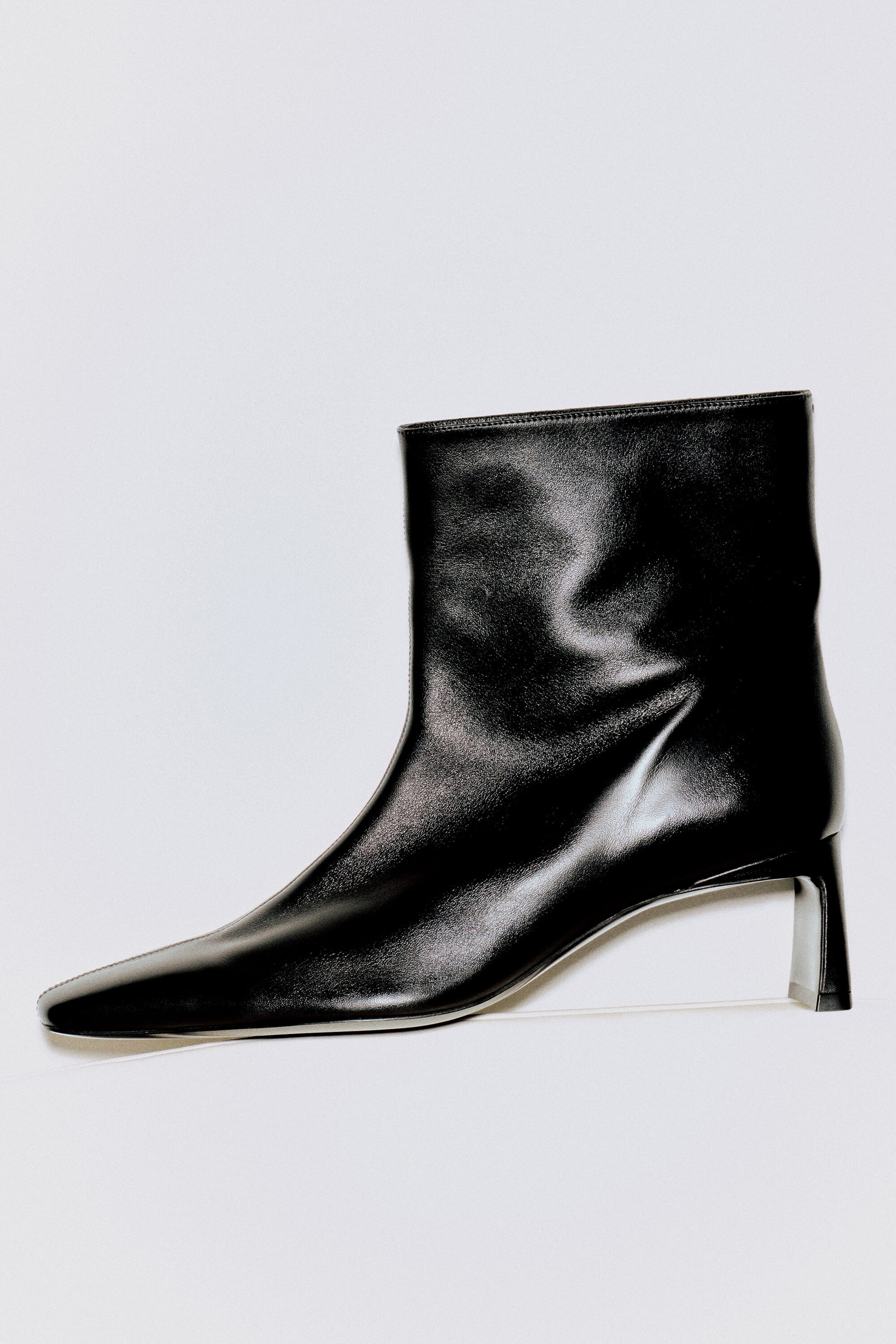 An angular leather heeled ankle boot is shown in a side profile facing left, featuring smooth black leather with an elongated square toe. The boot rests on a low, architectural block heel and rises to a straight, ankle-high shaft. An angular leather heeled ankle boot is shown in a side profile facing left, featuring smooth black leather with an elongated square toe. The boot rests on a low, architectural block heel and rises to a straight, ankle-high shaft.