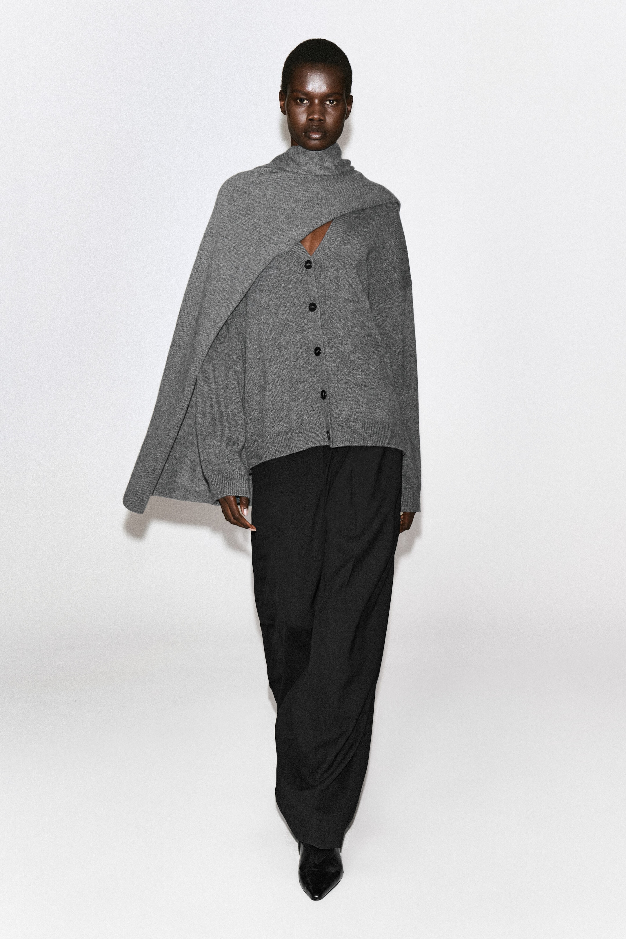 Un cardigan grigio in cashmere con scollo a V, bottoni frontali scuri e vestibilità rilassata è indossato dalla modella. Il cardigan è abbinato a una mantella in maglia grigia coordinata, drappeggiata sulla spalla, e a pantaloni neri a gamba larga. Un cardigan grigio in cashmere con scollo a V, bottoni frontali scuri e vestibilità rilassata è indossato dalla modella. Il cardigan è abbinato a una mantella in maglia grigia coordinata, drappeggiata sulla spalla, e a pantaloni neri a gamba larga.