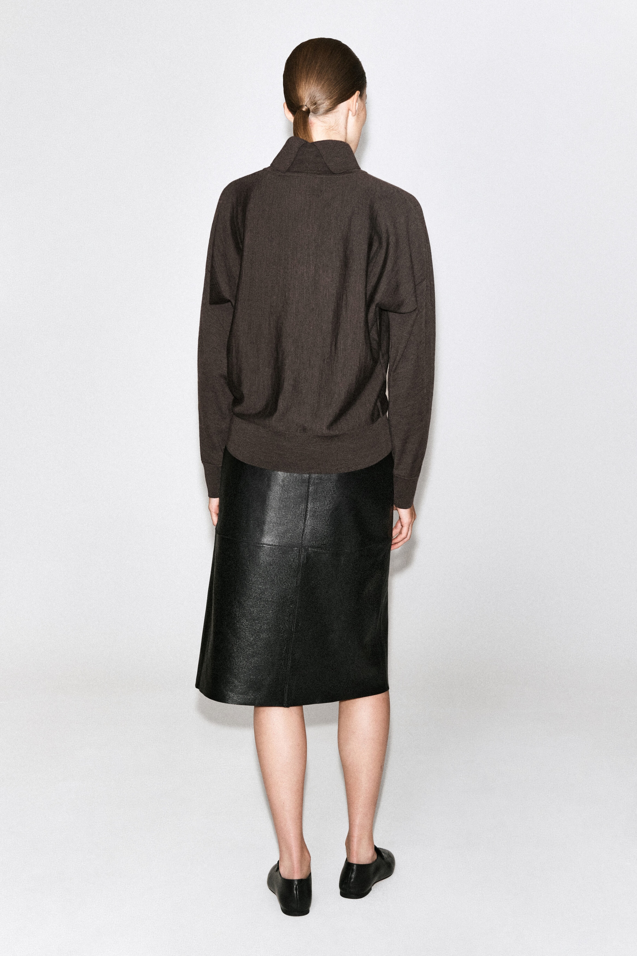 A dark brown, long-sleeved pullover sweater featuring a high funnel neck and ribbed trim is being worn by the model, who is viewed from behind. The sweater is styled with a black, knee-length leather skirt with panel detailing and black flat slip-on shoes. A dark brown, long-sleeved pullover sweater featuring a high funnel neck and ribbed trim is being worn by the model, who is viewed from behind. The sweater is styled with a black, knee-length leather skirt with panel detailing and black flat slip-on shoes.