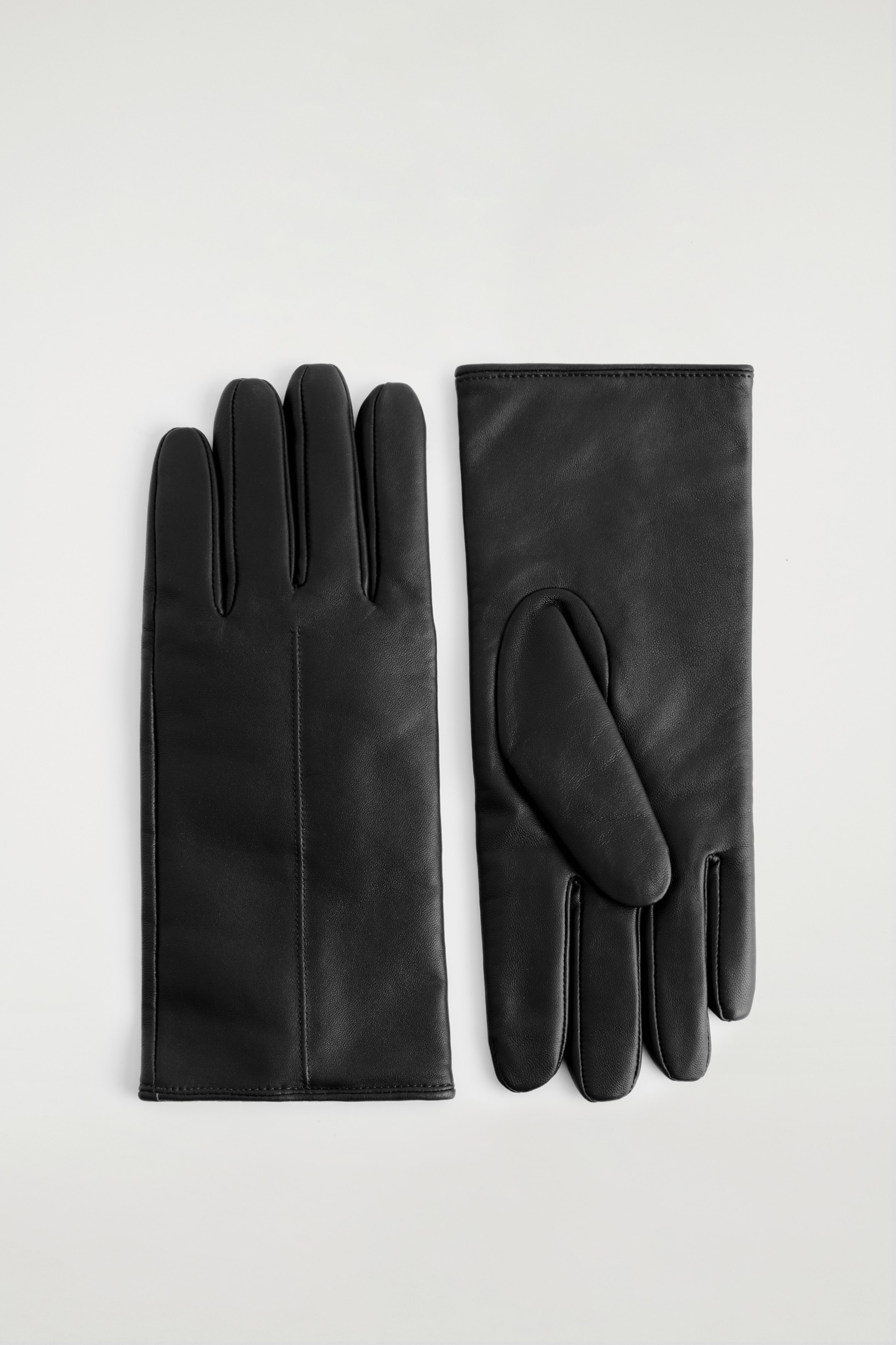 Grösseres Bild ansehen: Zwei schwarze Lederhandschuhe liegen flach, nebeneinander, aus einer Vogelperspektive. Der linke Handschuh zeigt seine Handfläche mit vertikalen Finger-Nähten, während der rechte Handschuh seinen Rücken zeigt, mit einem ovalen Daumenpanel und einer obergenähten Handgelenksmanschette.
