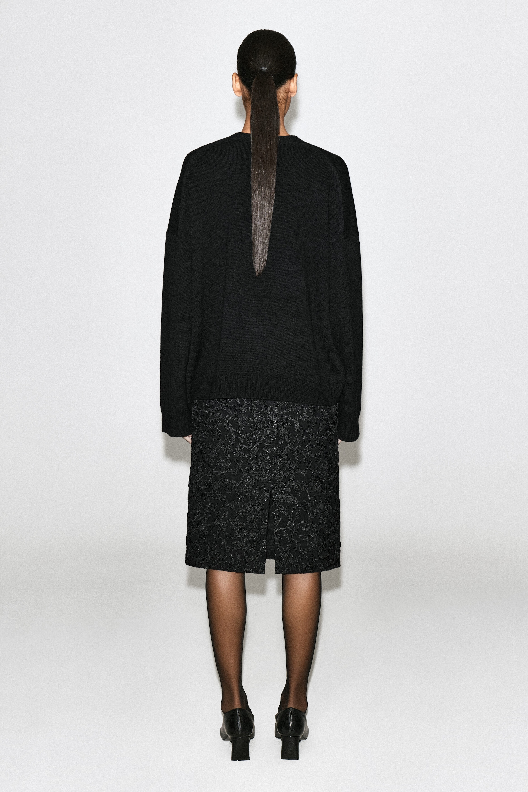 Un cardigan nero in maglia è mostrato dal retro, mettendo in risalto la sua vestibilità rilassata, le spalle scese e l’orlo a coste. Il cardigan è abbinato a una gonna nera fantasia al ginocchio per illustrarne la lunghezza e la caduta.