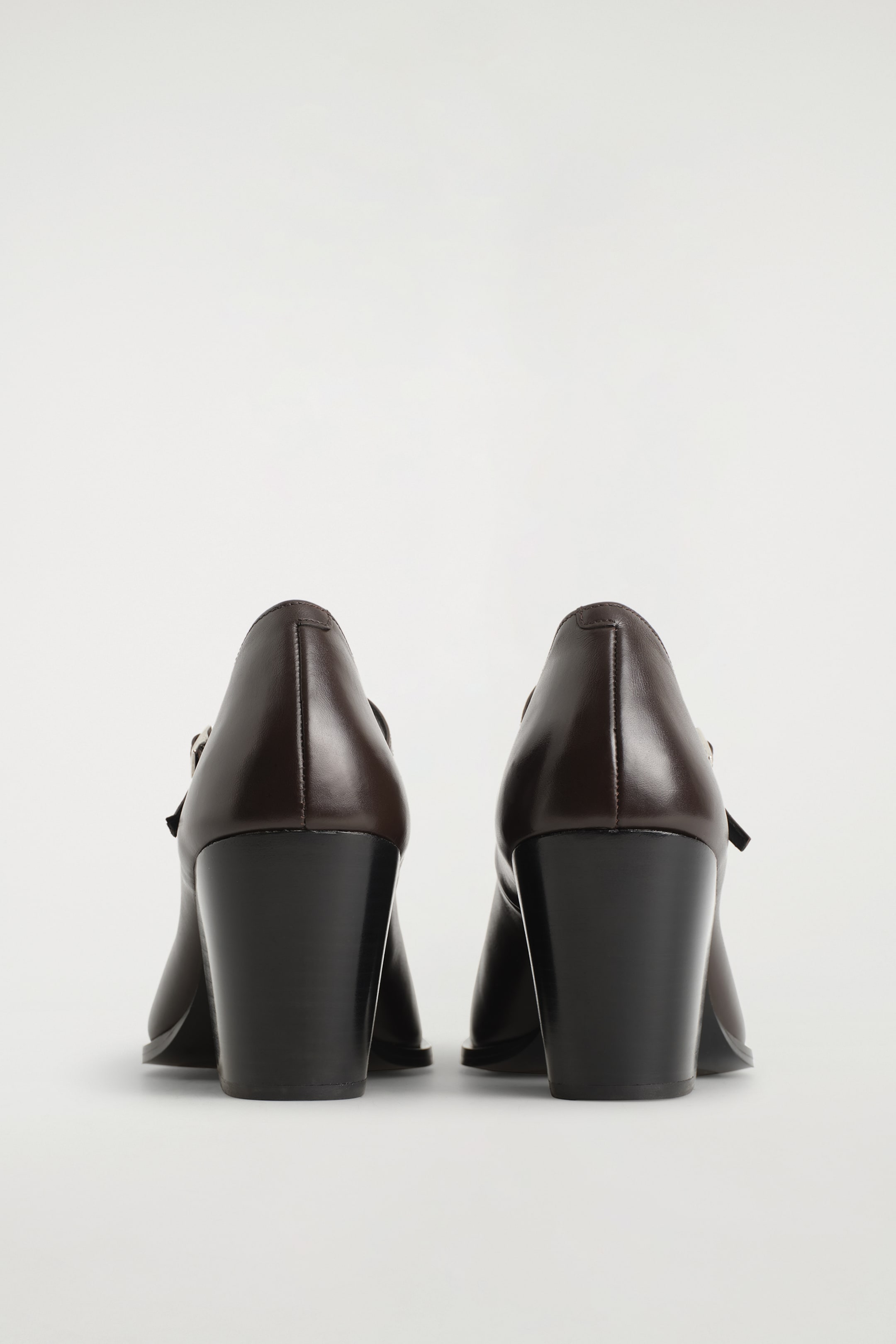 Agrandir l'image: Une paire de chaussures Mary Jane à talons en cuir marron foncé est présentée de l'arrière, mettant en valeur leur structure. Chaque chaussure est dotée d'un talon large et sombre ainsi que de coutures visibles le long de la couture arrière.