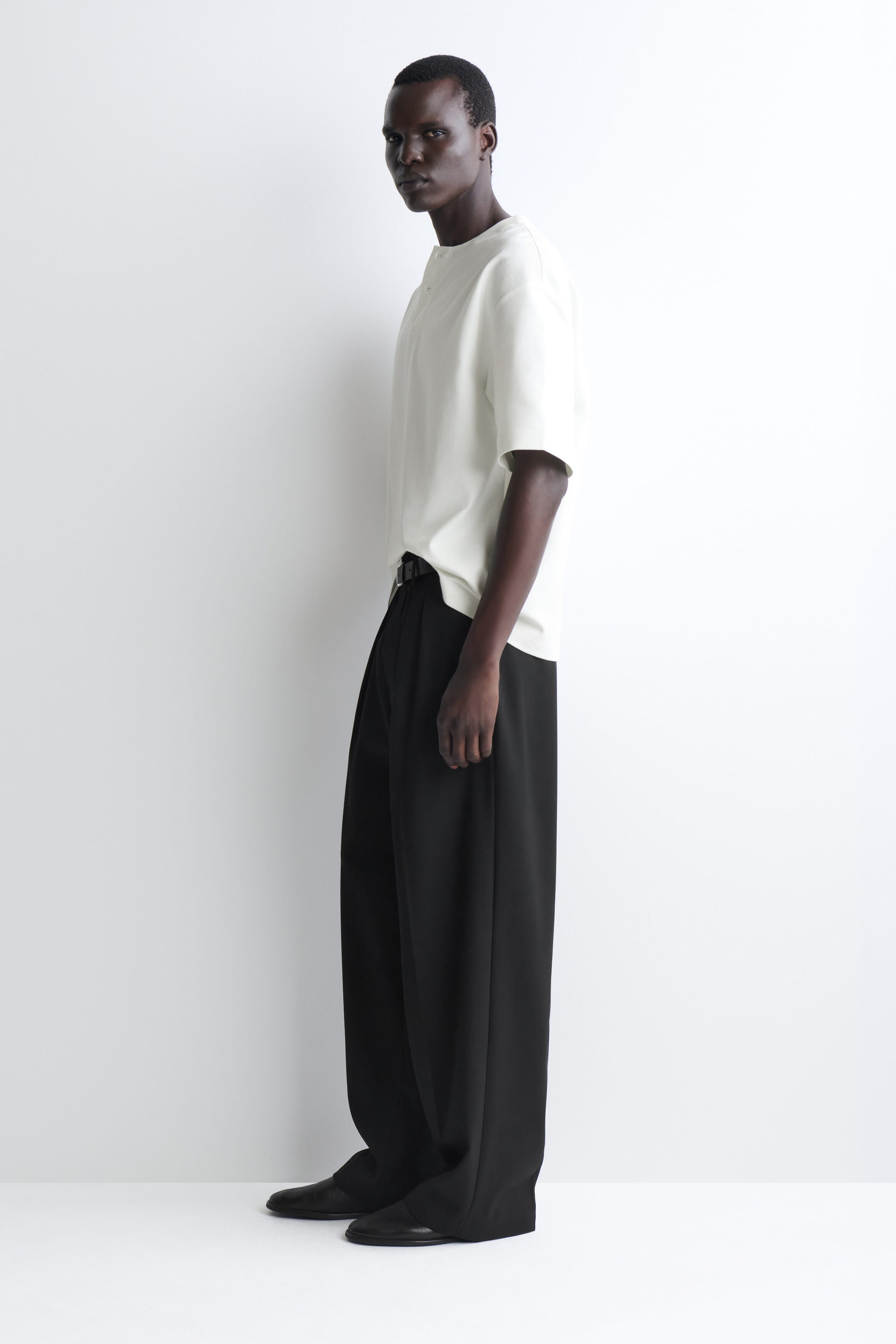 Le t-shirt henley oversize en jersey de coton blanc cassé présente une silhouette décontractée avec des manches courtes amples et un col boutonné minimaliste. Le t-shirt tombe librement sur un mannequin tourné vers la gauche, avec l’ourlet avant partiellement rentré dans un pantalon noir afin de mettre en valeur sa coupe fluide.