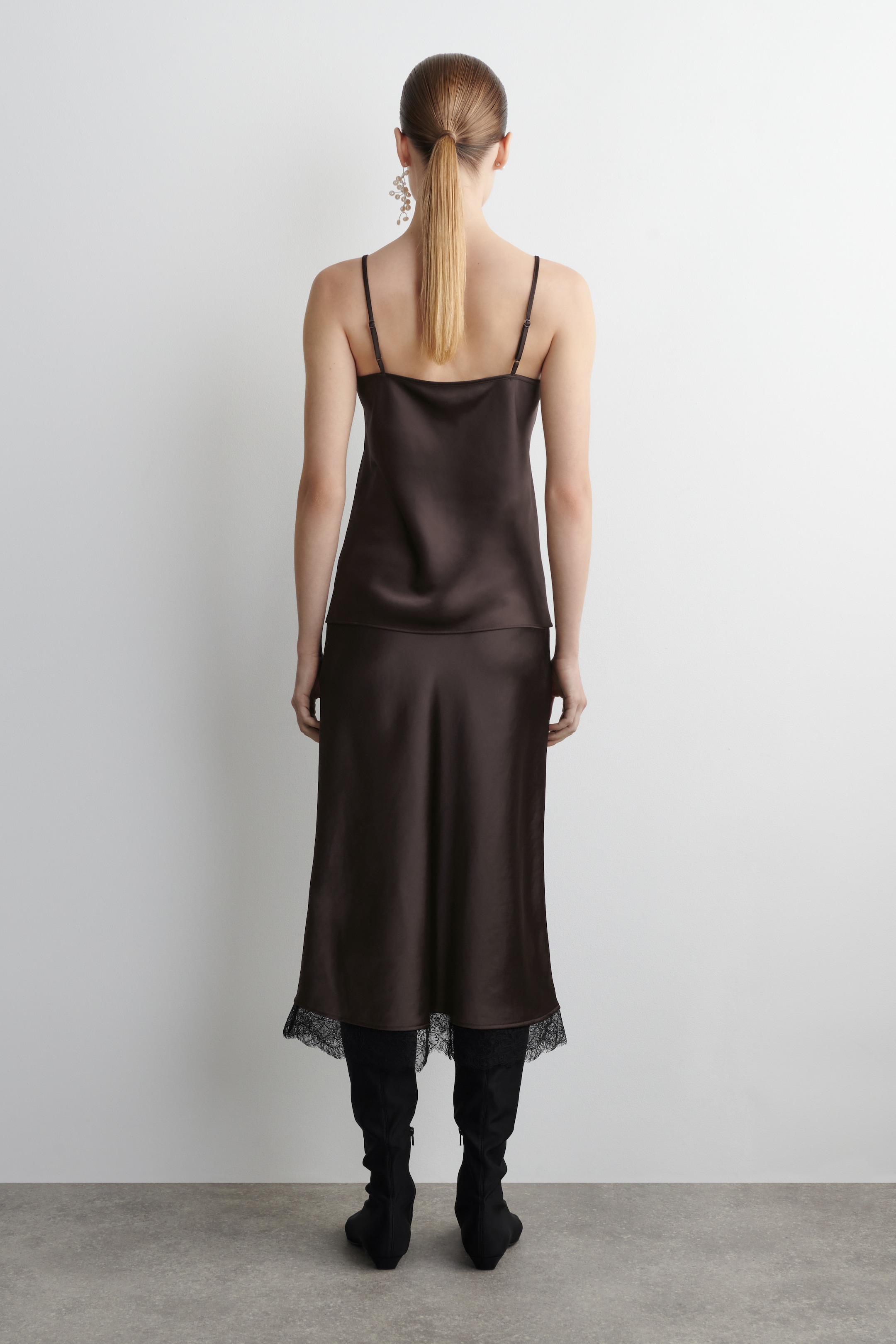 View larger image: A dark brown satin camisole top, featuring thin spaghetti straps, is being presented from the back. It is paired with a matching midi skirt that is displaying a black lace trim at its hem, creating a layered effect.