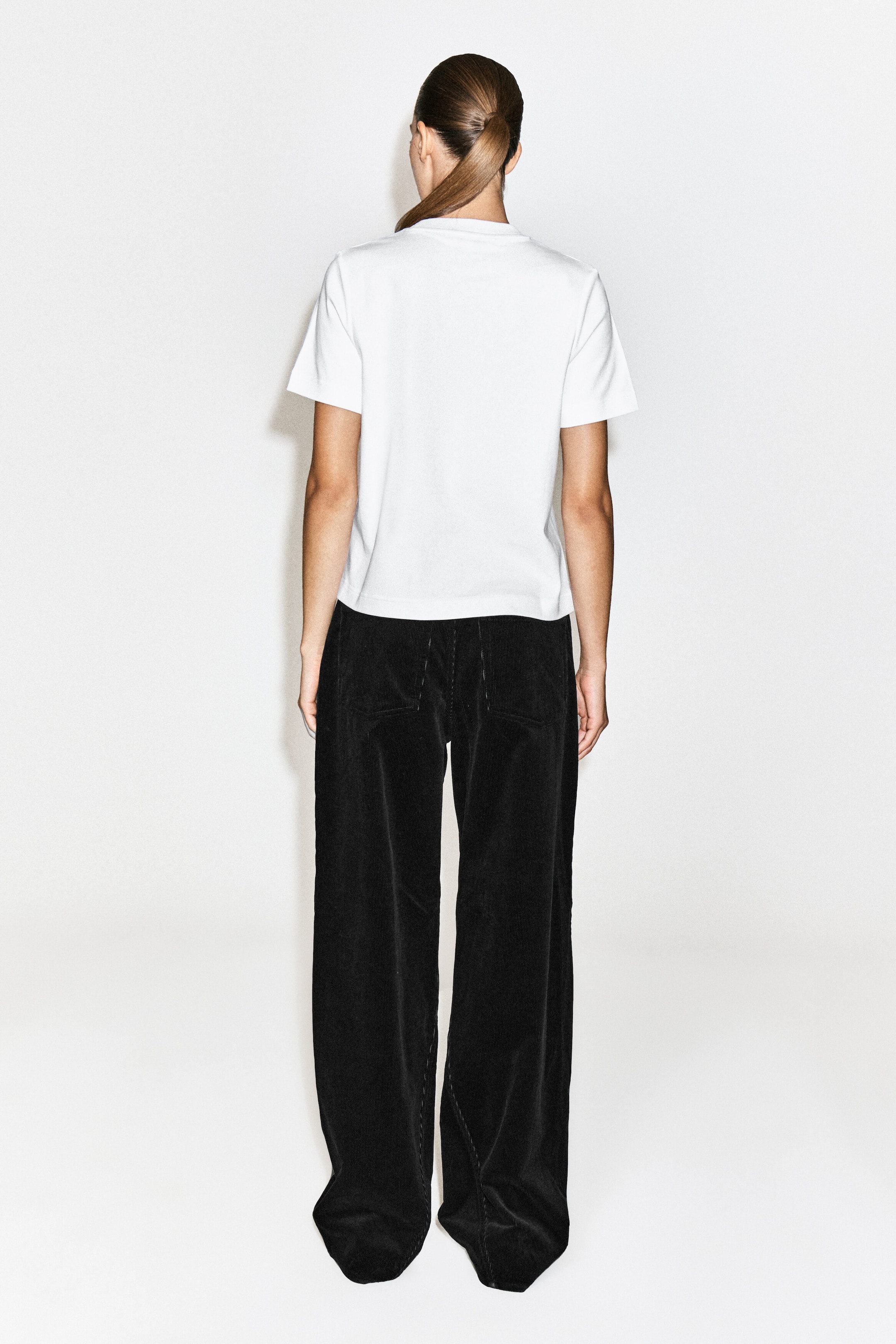 Black wide-leg trousers in a textured fabric are being worn by the model, paired with a white short-sleeved T-shirt. The trousers feature a relaxed fit, back pockets, and a long hem.
