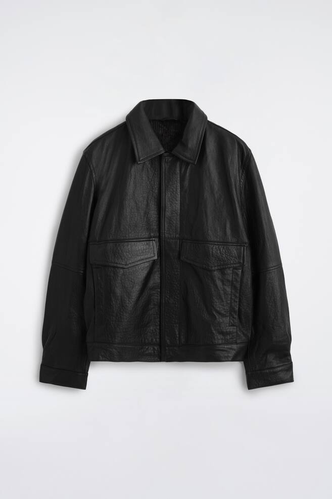 En svart flygjacka i lÃ¤der visas framifrÃ¥n och framhÃ¤ver sin strukturerade design och texturerade yta. Den har en klassisk spetsig krage, dold stÃ¤ngning framtill och tvÃ¥ stora brÃ¶stfickor med lock. - 1