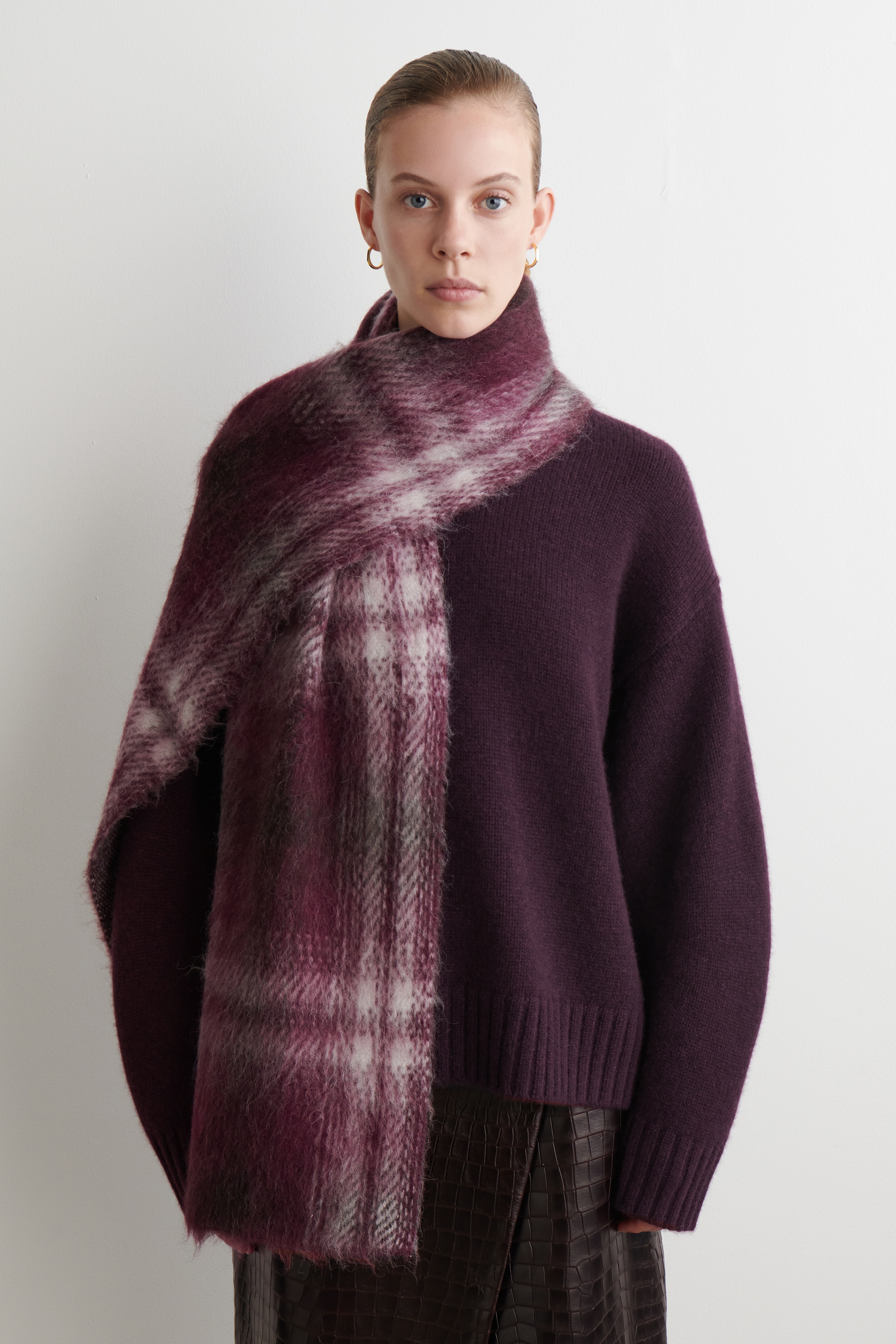 Agrandir l'image: Un pull col montant prune profond, avec des poignets et un ourlet côtelés, est porté face vers l'avant. Une écharpe à carreaux pelucheuse, affichant des nuances de violet, de blanc et de gris, est drapée autour du cou et sur les épaules, avec une extrémité pendante sur le devant.