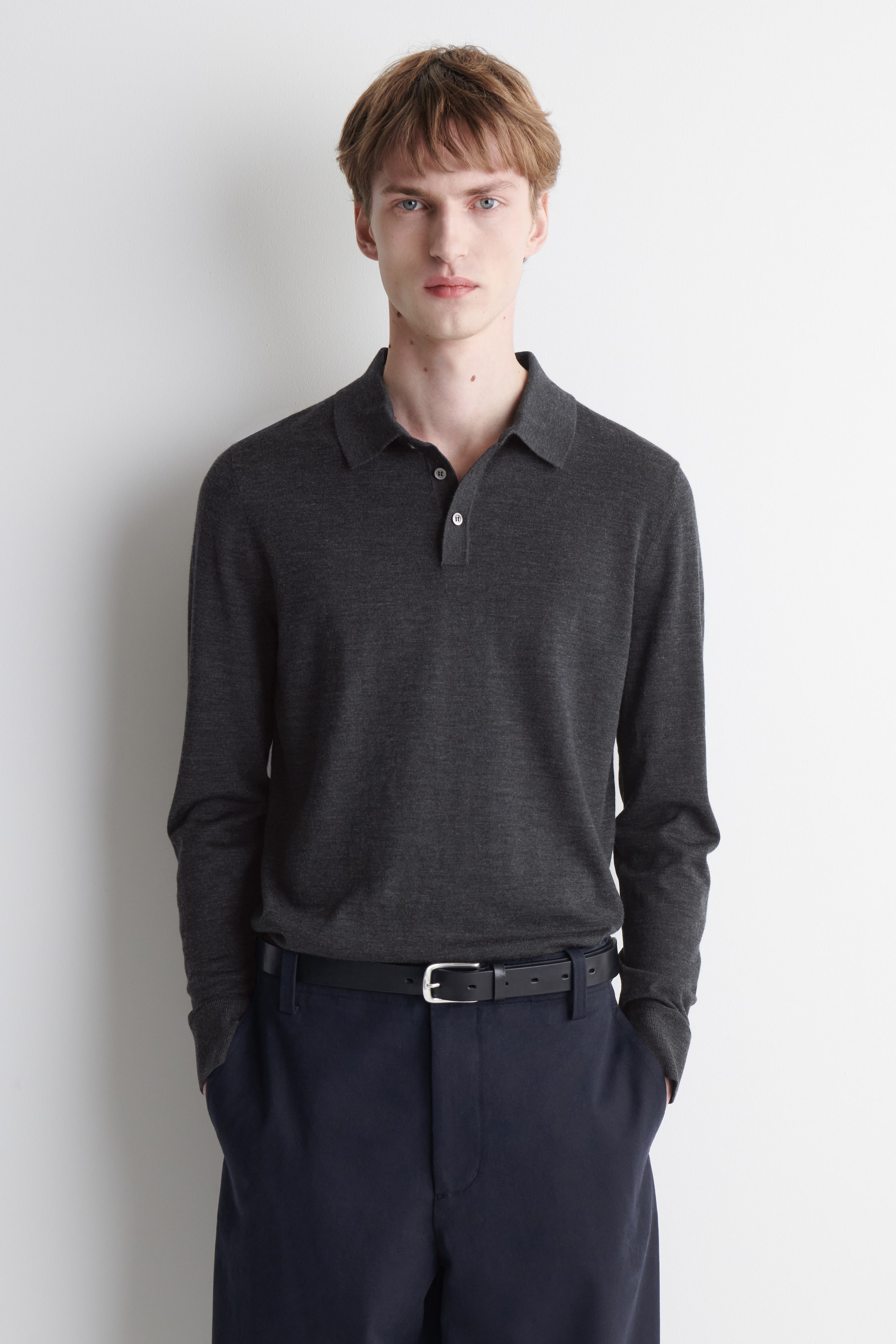 View larger image: A dark grey long-sleeved knitted polo shirt is being worn by the model, featuring a polo collar and a two-button placket with light-coloured buttons. The shirt is styled with dark blue trousers and a black belt.