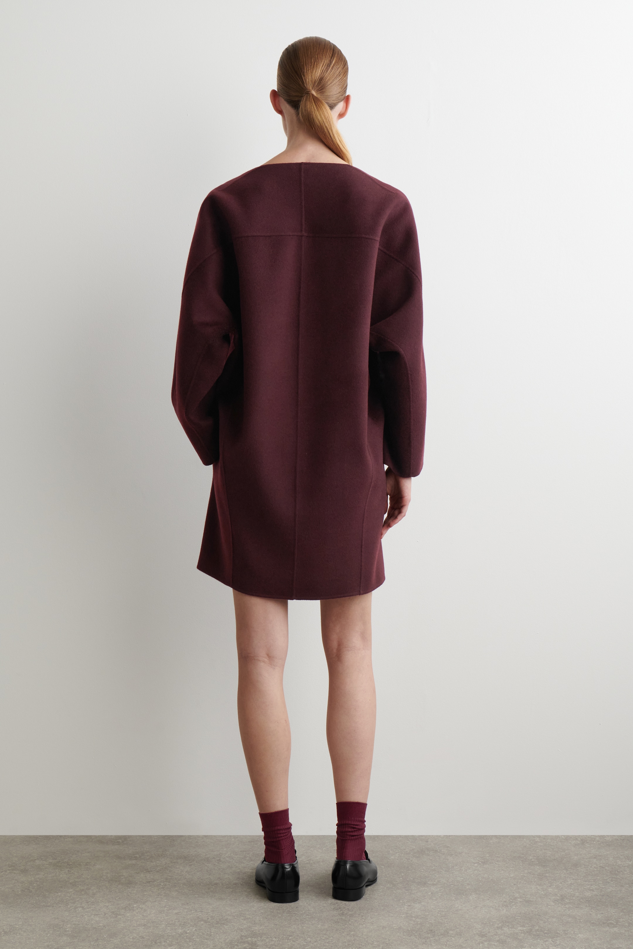 Agrandir l'image: La ROBE MINI EN LAINE DOUBLE FACE est vue de dos, arborant une couleur bordeaux profond et une coupe ample et droite. Elle est conçue avec des manches larges et longues, un large décolleté en forme de cuillère, et des coutures horizontales et verticales proéminentes définissant sa structure sur le haut du dos et le long du centre.
