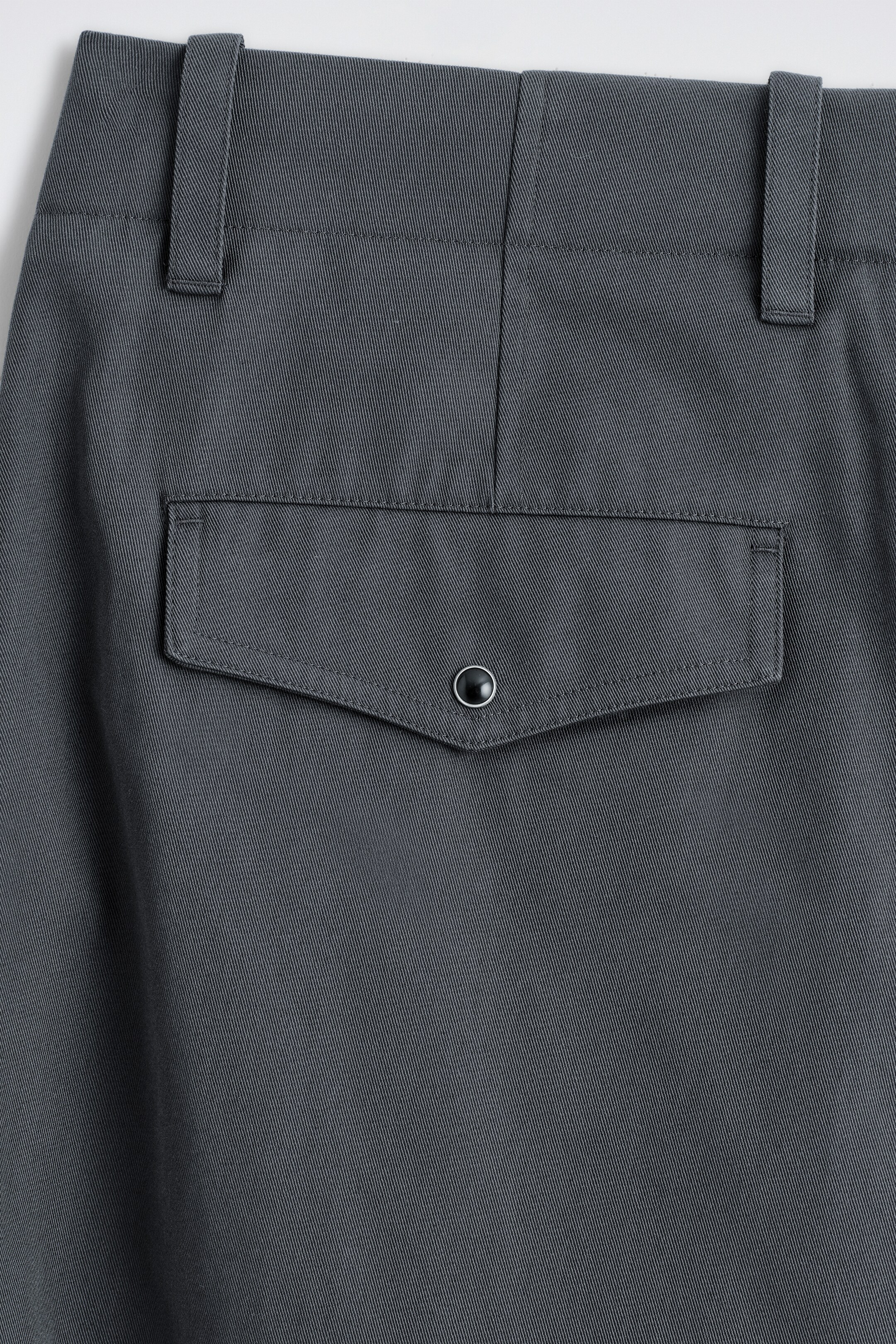 View larger image: The back of dark gray pants is featuring a twill weave fabric, with a waistband and two visible belt loops. A single flap pocket is centrally positioned below the waistband, showcasing a pointed bottom edge and a black snap button with a metallic rim securing the flap.