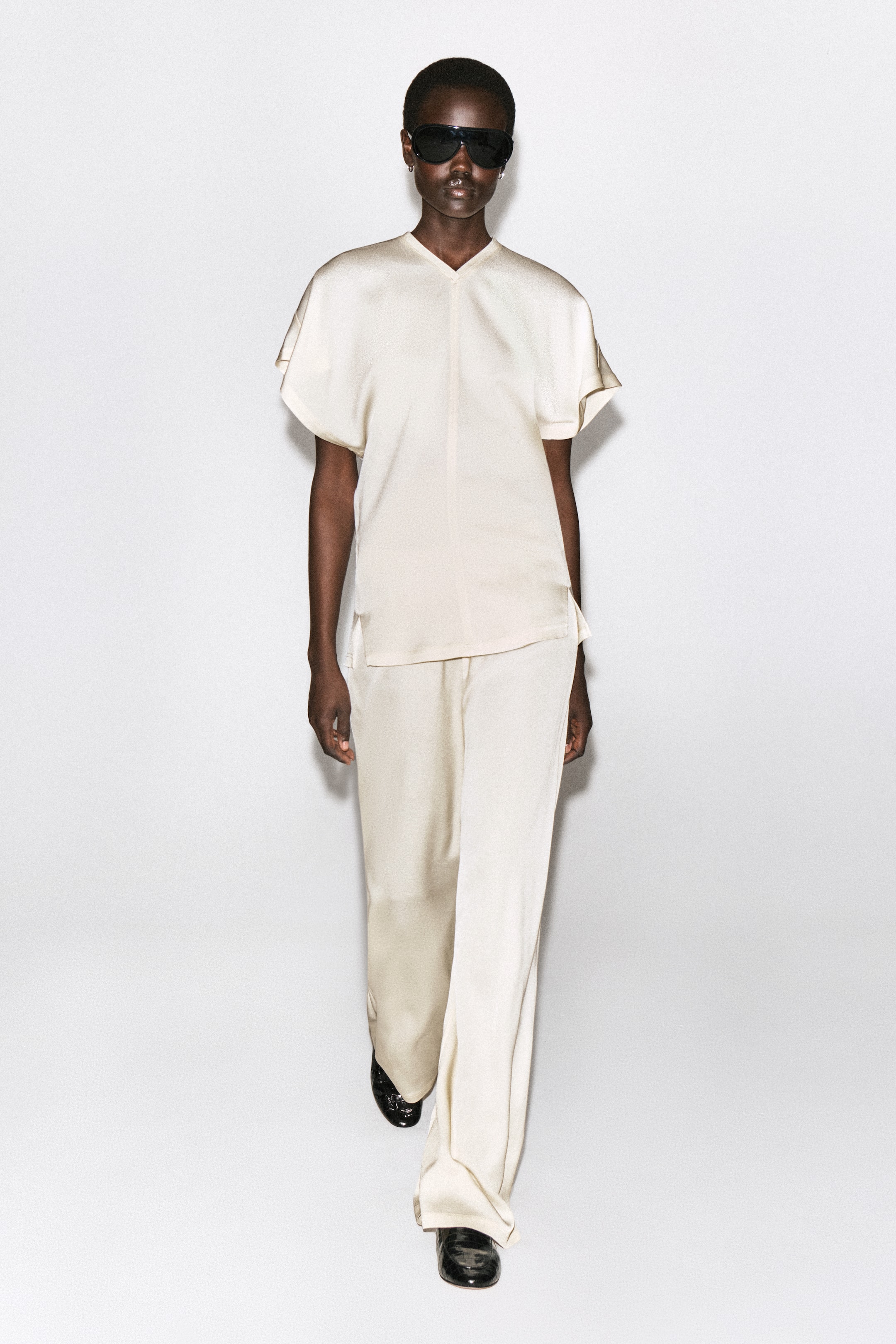 Grösseres Bild ansehen: Die elastischen Crêpe-Hosen mit geradem Bein in Creme fallen in einer lockeren, fliessenden Silhouette in der Vorderansicht. Sie werden mit einem passenden kurzärmeligen Oberteil in Creme kombiniert, das den Bund bedeckt, wobei die langen Säume leicht über schwarzen Schuhen aufliegen.