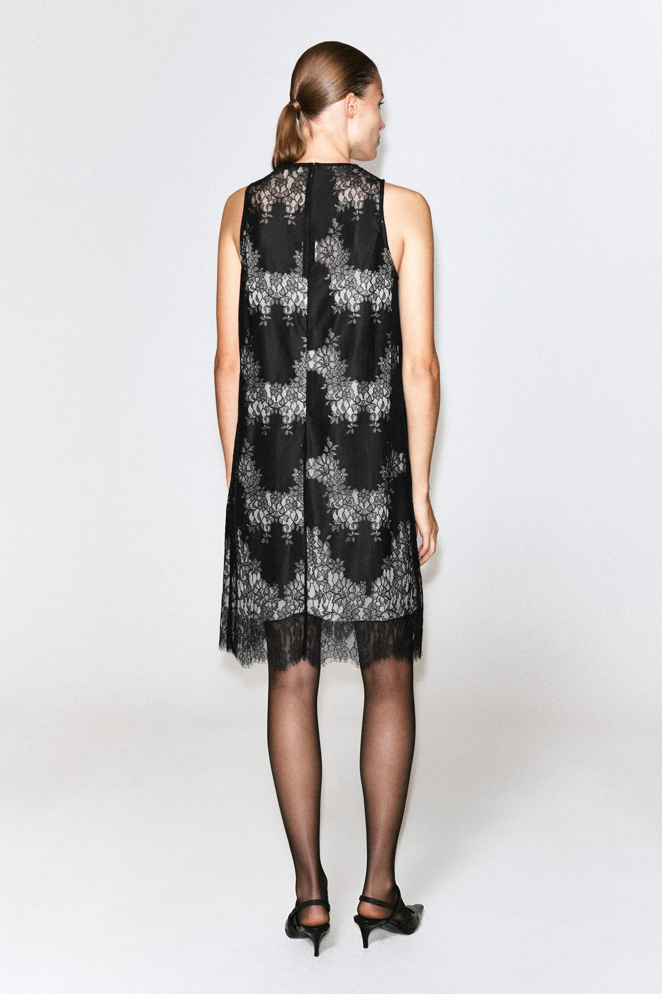 A sleeveless lace midi dress featuring a black floral lace overlay and a contrasting light underlay is being worn by the model. The straight-cut dress features a high neckline, a central back seam and a scalloped eyelash lace hem, and is being styled with sheer black tights and black pointed-toe slingback heels. A sleeveless lace midi dress featuring a black floral lace overlay and a contrasting light underlay is being worn by the model. The straight-cut dress features a high neckline, a central back seam and a scalloped eyelash lace hem, and is being styled with sheer black tights and black pointed-toe slingback heels.