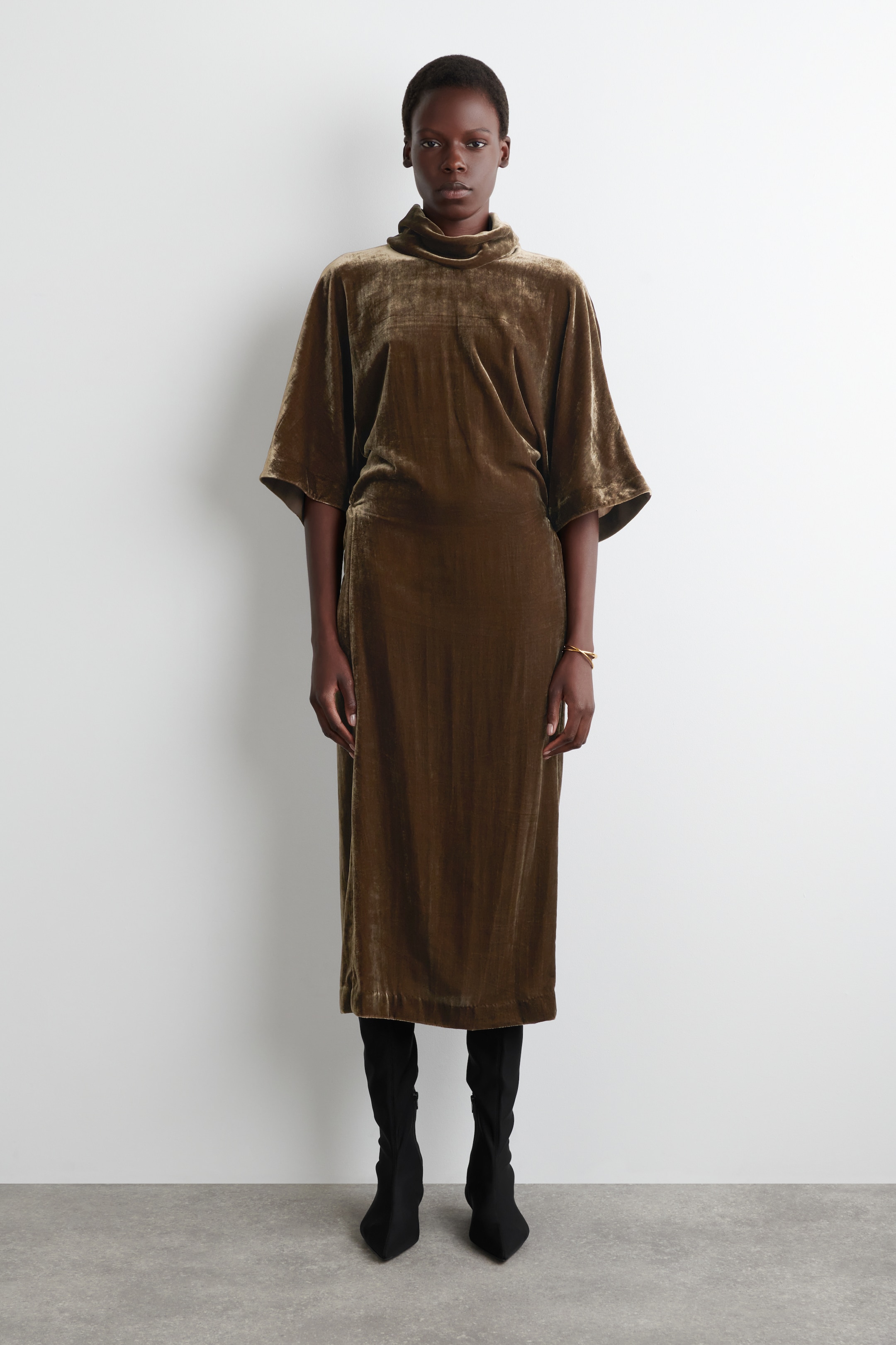 Grösseres Bild ansehen: Das olivgrüne Kleid aus zerdrücktem Samt ist nach vorne gerichtet und verfügt über einen drapierten Wasserfallausschnitt sowie breite, ellbogenlange Dolman-Ärmel. Es präsentiert eine lockere Passform, die locker durch das Oberteil fällt und in einen geraden Midirock übergeht.