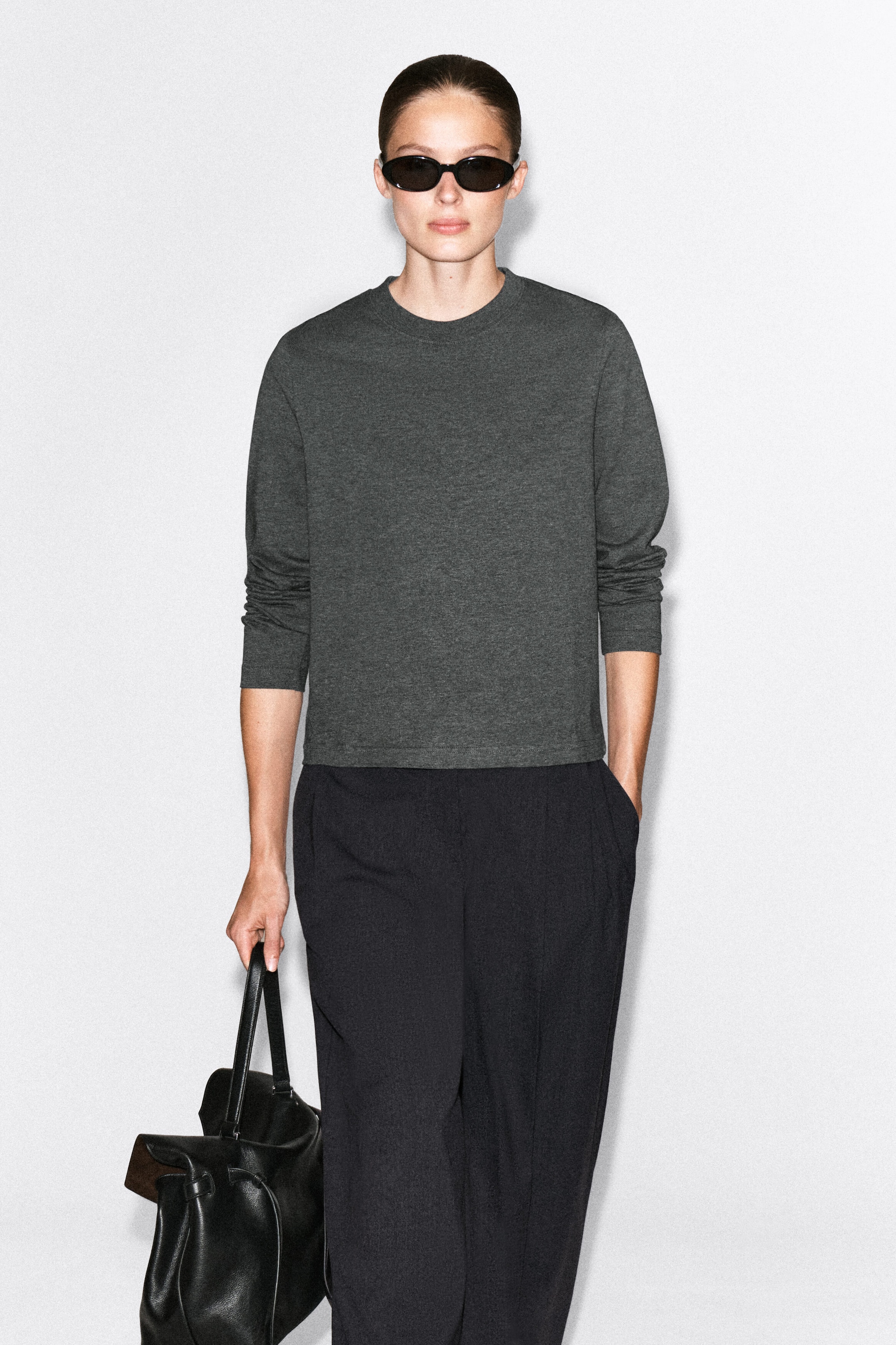 The model is wearing a dark gray long-sleeve T-shirt with a crew neckline and straight hem. It is styled with dark trousers and a black leather handbag held in one hand.