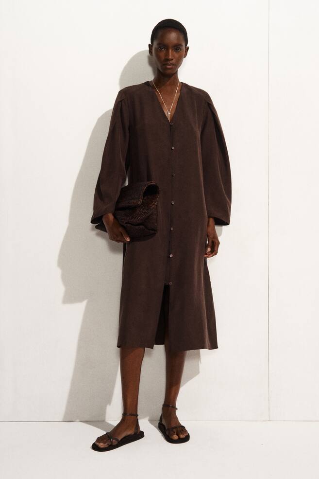 Une robe midi marron foncé est présentée de face, avec un décolleté en V et une patte de boutonnage pleine longueur. Elle est conçue avec des manches longues volumineuses froncées aux épaules et tombe dans une silhouette droite jusqu'en dessous des genoux, avec les boutons du bas laissés ouverts formant une fente à l'avant. - 1