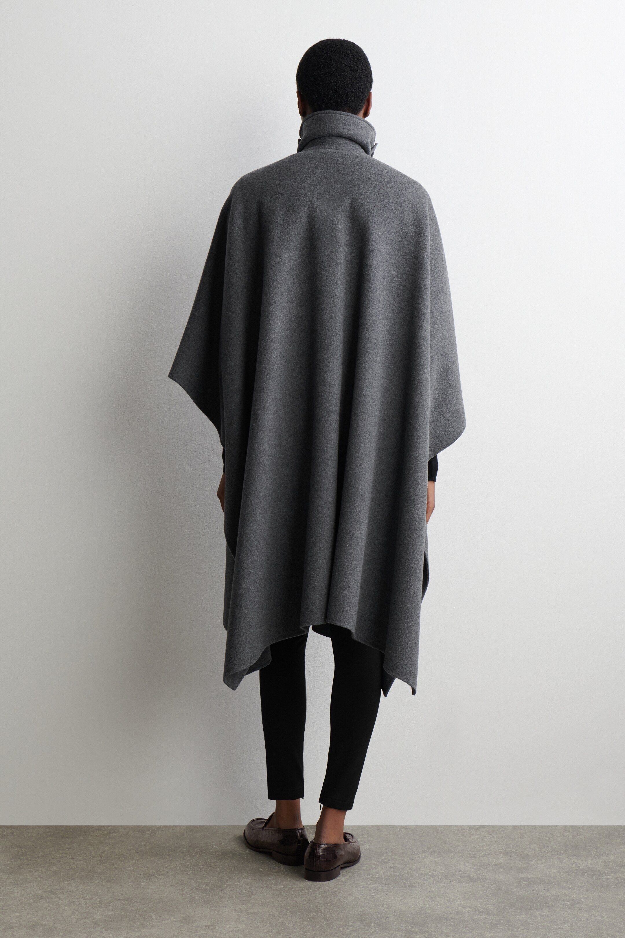 Agrandir l'image: Un poncho gris foncé est présenté de dos, mettant en valeur sa draperie ample et fluide ainsi qu'un ourlet asymétrique. Il est doté d'un col montant avec des boutons visibles et est superposé sur un haut noir à manches longues et des leggings noirs, avec des mocassins marron foncé pour compléter le look.