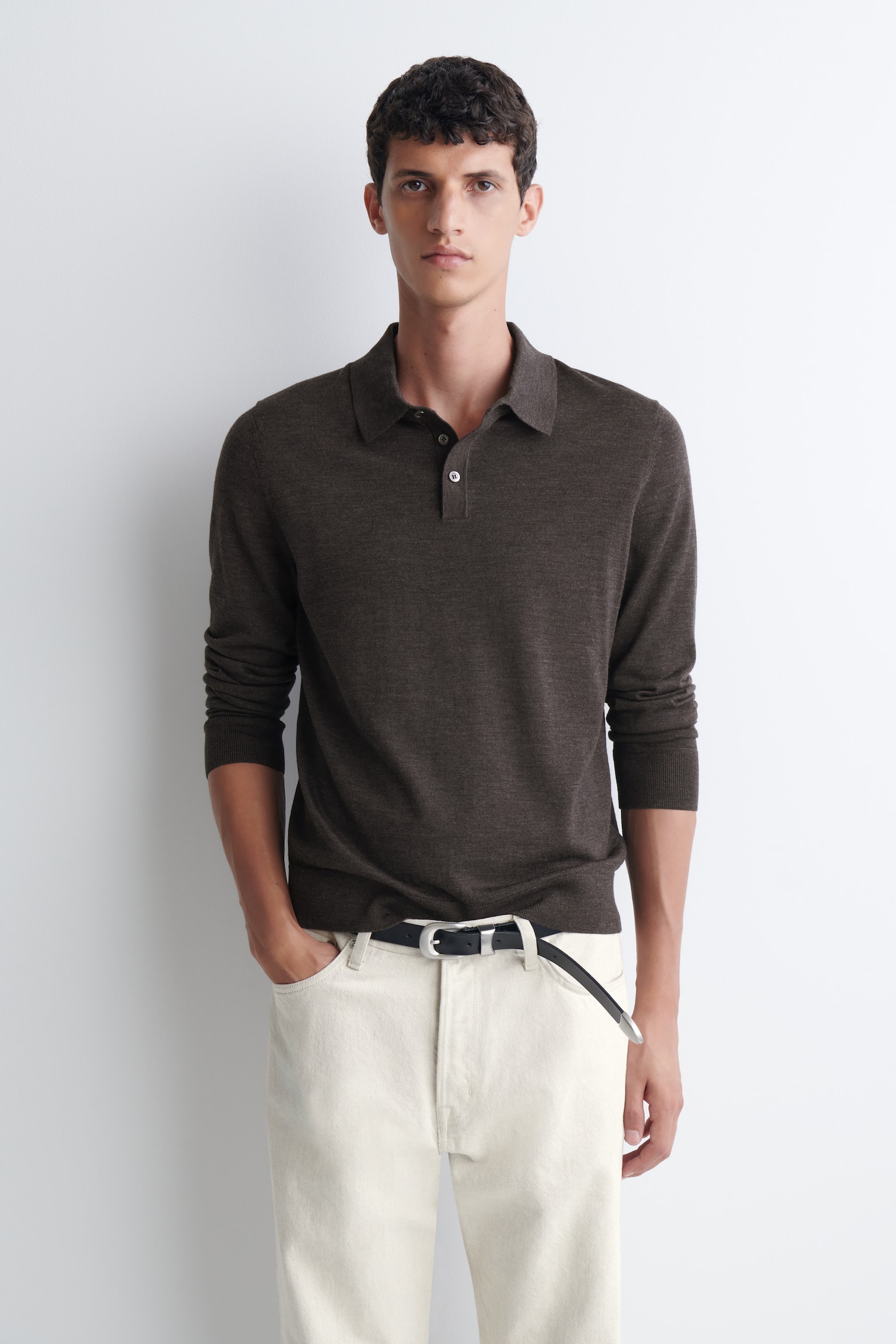 Der Model trägt einen dunkelbraunen Strick-Pullover im Polostil mit langen Ãrmeln, kombiniert mit einer cremefarbenen Hose und einem schwarzen Ledergürtel. Der Pullover verfügt über einen klassischen Kragen, eine Dreiknopfleiste und gerippte Bündchen, die leicht bis zu den Unterarmen hochgeschoben sind. - 1