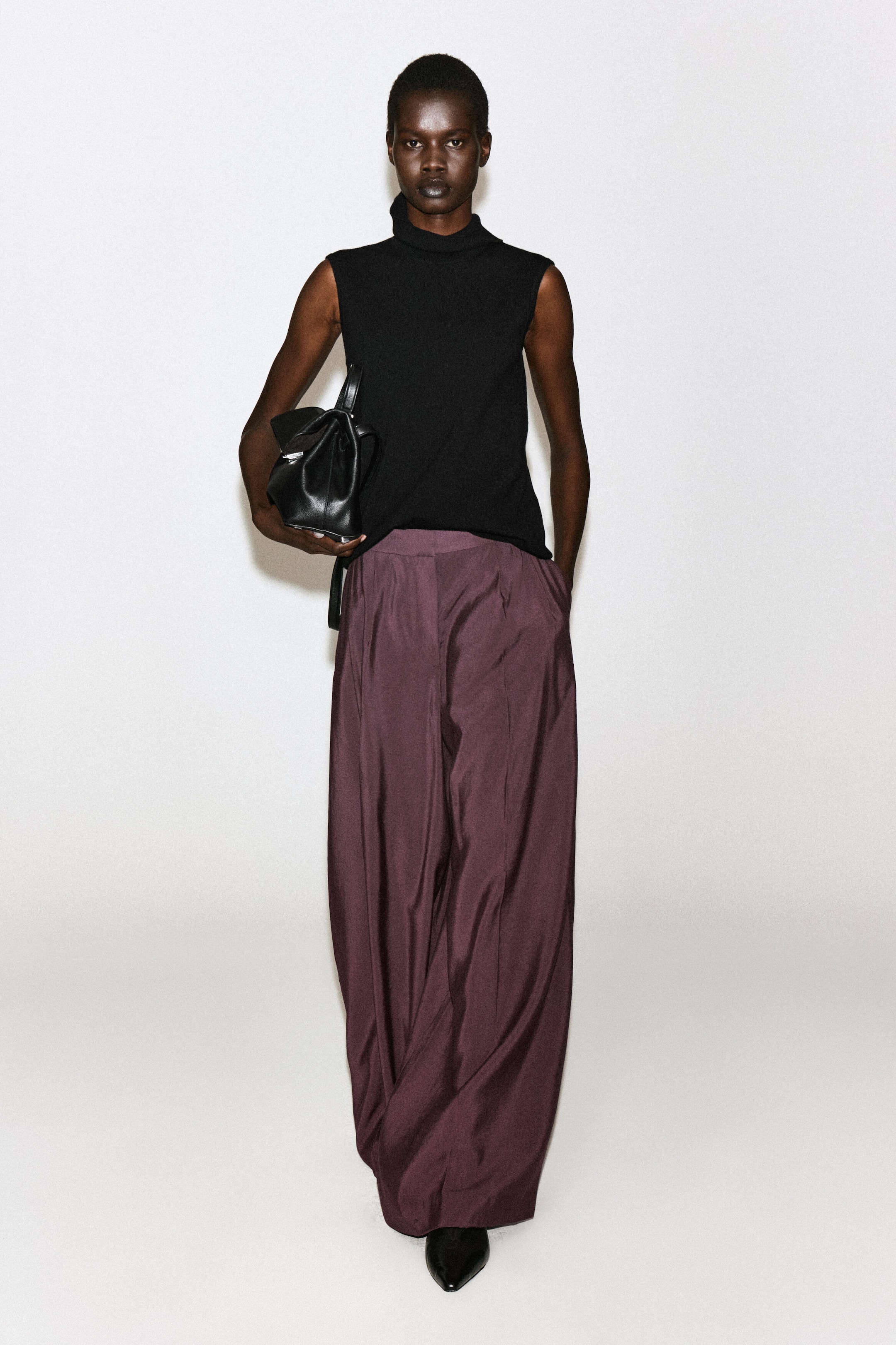 Plissierte Wide-Leg-Hose in einem dunklen Pflaumenton, vom Model getragen, das mit einer Hand in der Tasche steht. Die hoch geschnittene Hose überzeugt mit fließendem Fall und vorderen Bügelfalten und wird mit einem schwarzen ärmellosen Top mit Stehkragen sowie einer schwarzen Ledertasche kombiniert. Plissierte Wide-Leg-Hose in einem dunklen Pflaumenton, vom Model getragen, das mit einer Hand in der Tasche steht. Die hoch geschnittene Hose überzeugt mit fließendem Fall und vorderen Bügelfalten und wird mit einem schwarzen ärmellosen Top mit Stehkragen sowie einer schwarzen Ledertasche kombiniert.