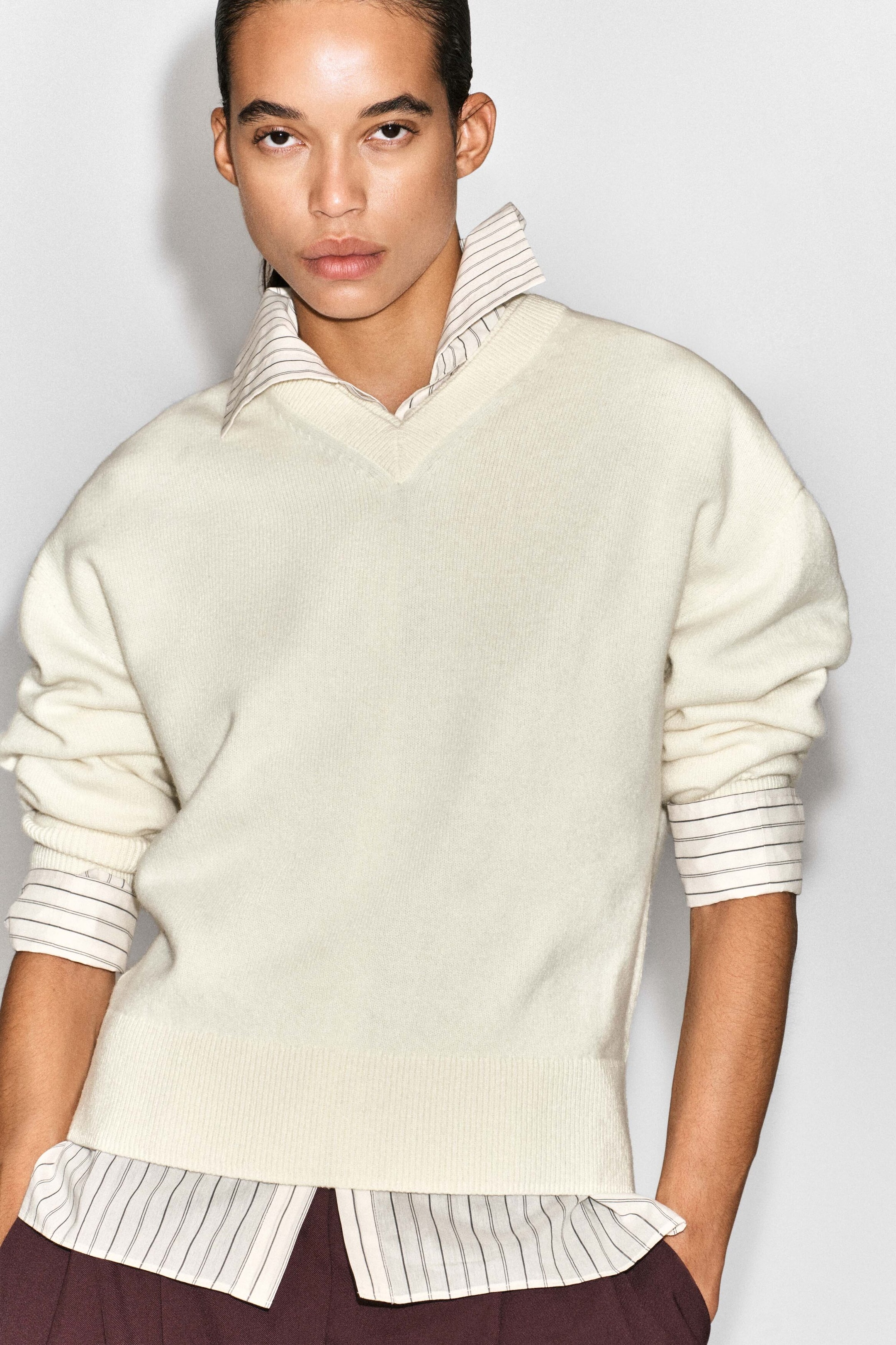 The cream-coloured merino wool V-neck jumper is shown from the front, featuring a relaxed fit with a ribbed neckline, cuffs, and hem. It is layered over a white pinstriped collared shirt, with the shirt's collar, cuffs, and lower hem visible. The cream-coloured merino wool V-neck jumper is shown from the front, featuring a relaxed fit with a ribbed neckline, cuffs, and hem. It is layered over a white pinstriped collared shirt, with the shirt's collar, cuffs, and lower hem visible.