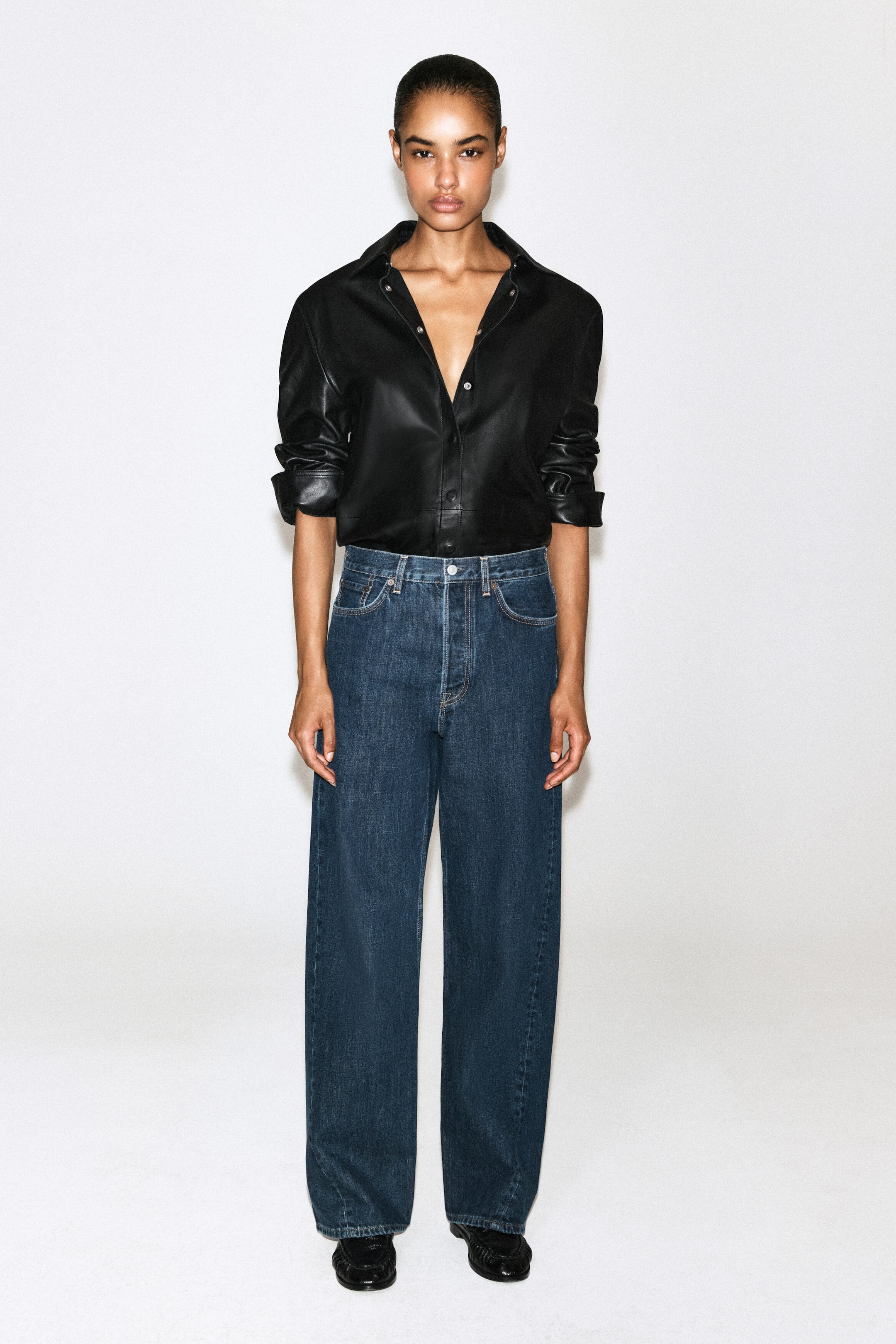 A model is standing facing forward, wearing high-waisted, wide-leg jeans in a dark blue wash with classic five-pocket styling. The jeans are paired with a tucked-in black leather button-down shirt with sleeves rolled up to the elbows. A model is standing facing forward, wearing high-waisted, wide-leg jeans in a dark blue wash with classic five-pocket styling. The jeans are paired with a tucked-in black leather button-down shirt with sleeves rolled up to the elbows.