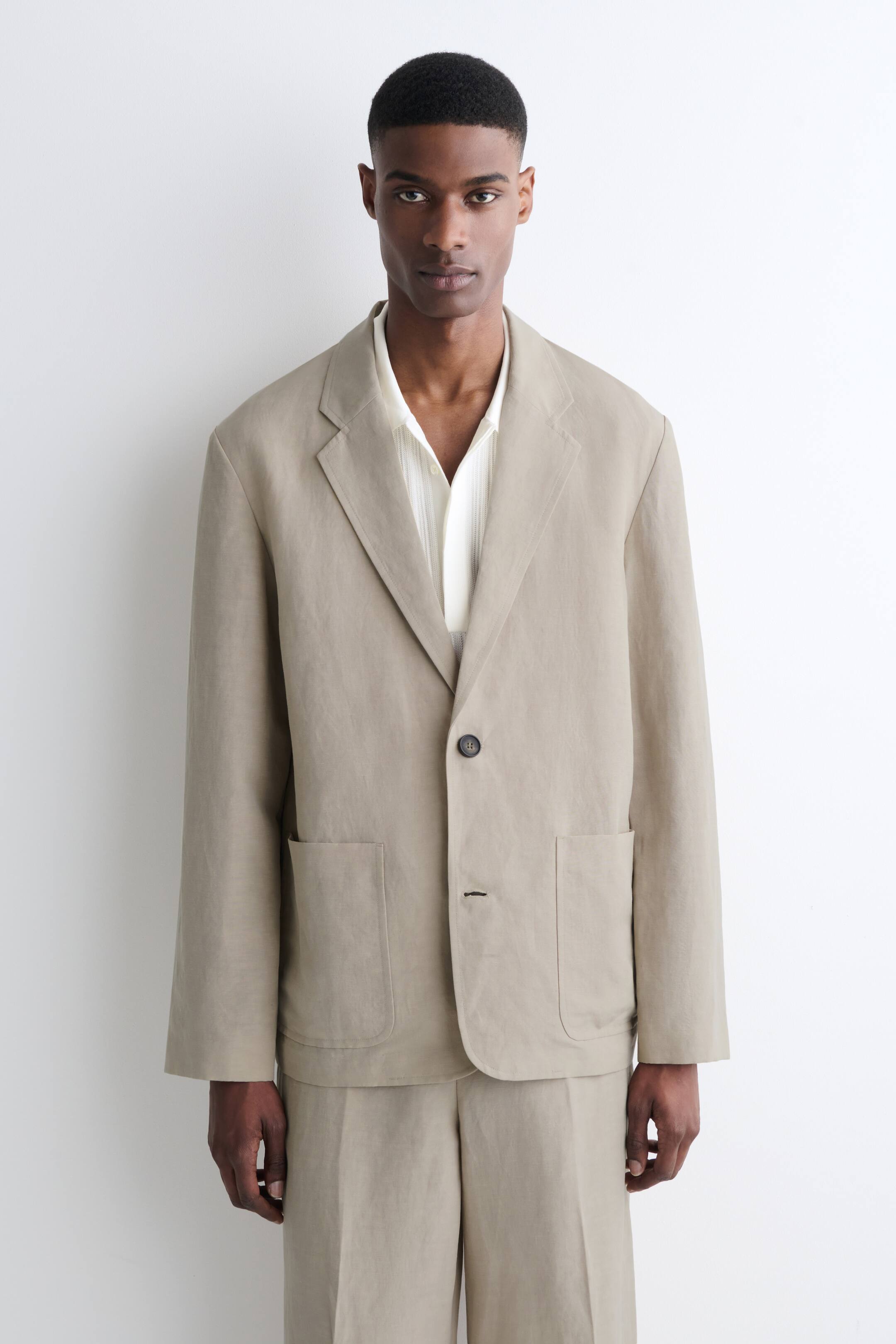 Visualizza immagine più grande: "Una giacca beige chiaro effetto lino è presentata frontalmente, caratterizzata da revers a dente, una chiusura con un singolo bottone scuro e due grandi tasche applicate arrotondate sui fianchi, abbinata a una camicia leggera dalla texture simile e pantaloni coordinati."