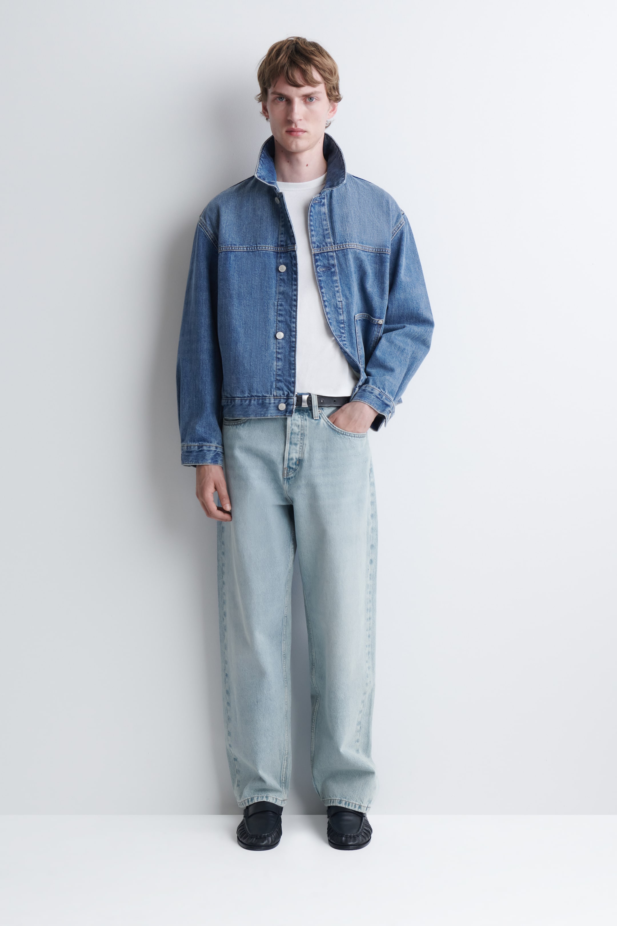 Visa större bild: Modellen bär en blå jeansjacka och ljusa, vida jeans över en vit t-shirt. Jackan har en hög, upprätt krage och knäppning med knappar, och modellen står med ena handen vilande i jeansens framficka.