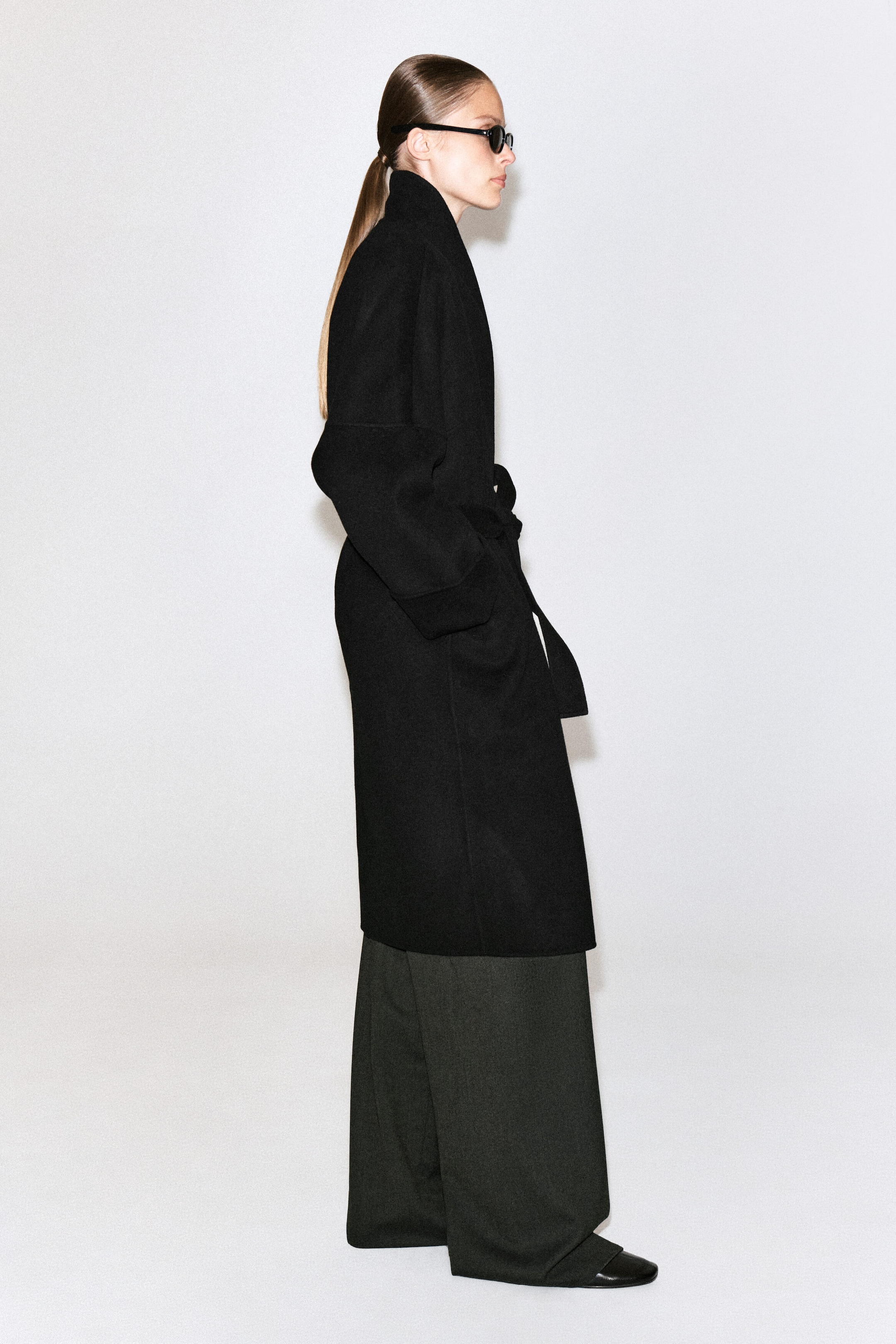 Un manteau-shawl oversize en laine double face noir est porté par le mannequin en profil latéral. Le manteau présente un col montant, des épaules tombantes et une ceinture à nouer à la taille, porté sur un pantalon large sombre. Un manteau-shawl oversize en laine double face noir est porté par le mannequin en profil latéral. Le manteau présente un col montant, des épaules tombantes et une ceinture à nouer à la taille, porté sur un pantalon large sombre.