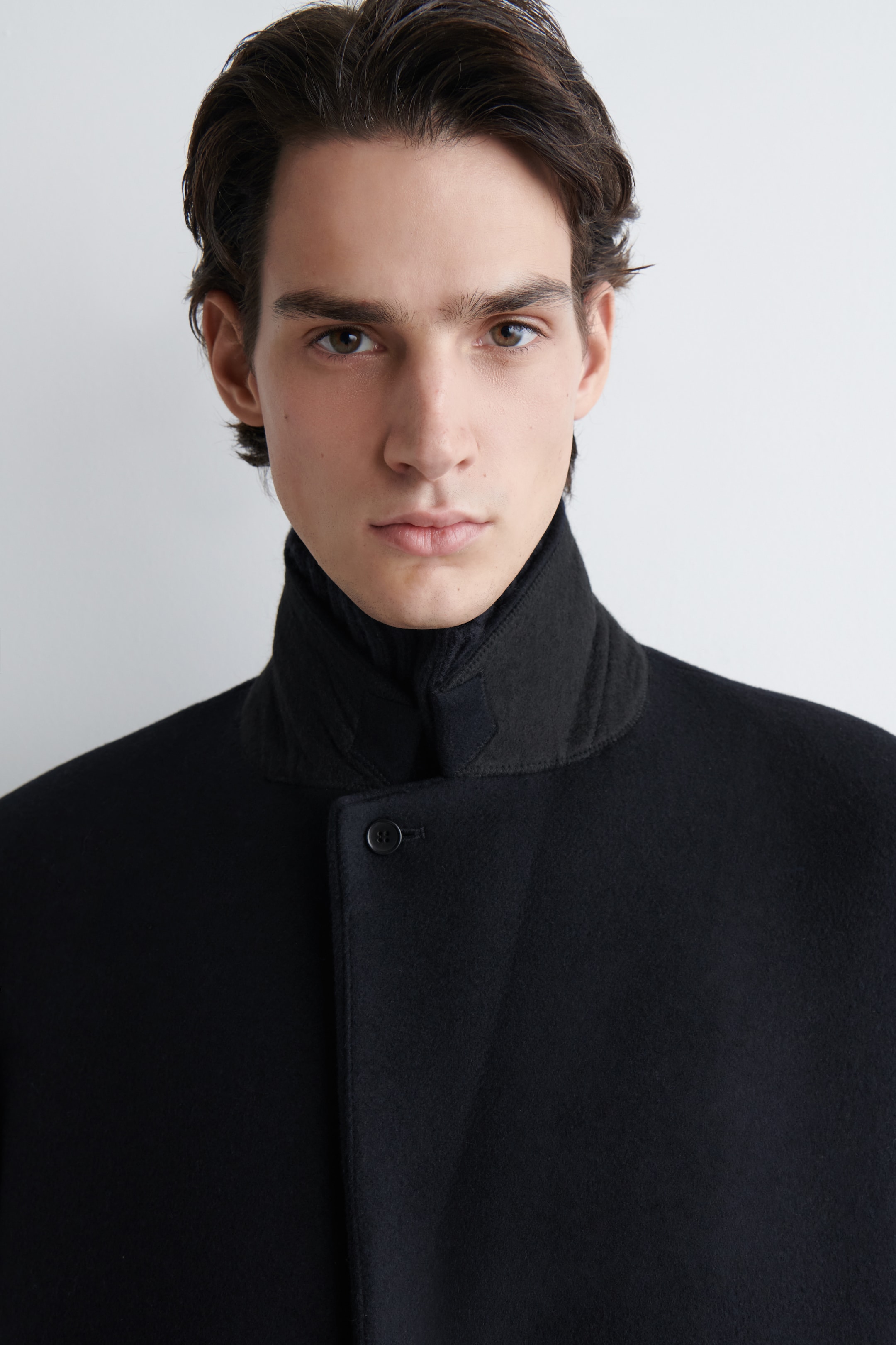 View larger image: A black felted coat is being shown from a frontal perspective, featuring a stand collar and a visible black button closure on its front. A dark, ribbed high-neck garment is being layered underneath the coat's collar.