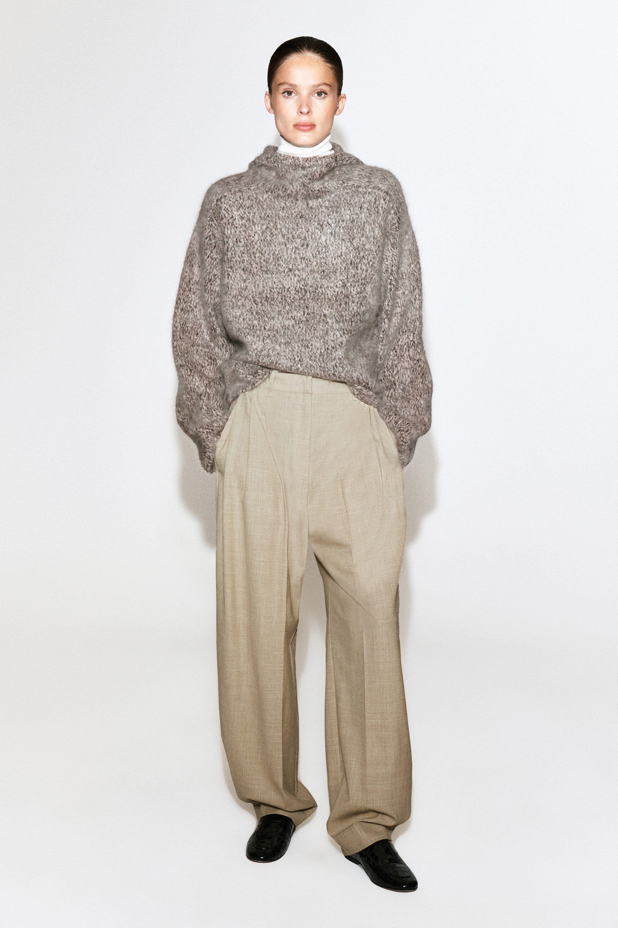 A chunky brushed cashmere hoodie in a textured grey-brown mélange knit is being worn by the model. The relaxed-fit garment features a high collar and is layered over a white high-neck top and paired with beige wide-leg trousers. A chunky brushed cashmere hoodie in a textured grey-brown mélange knit is being worn by the model. The relaxed-fit garment features a high collar and is layered over a white high-neck top and paired with beige wide-leg trousers.