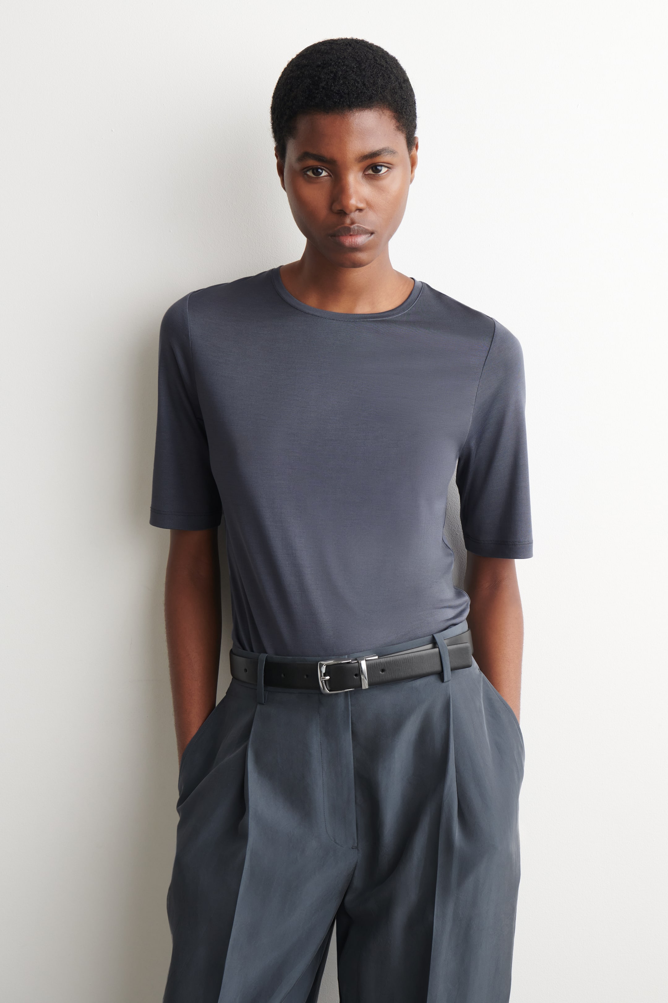 Se større billede: En stålblå T-shirt med rund hals og korte ærmer bæres af en model. T-shirten har en afslappet pasform og er stukket ned i mørkegrå bukser, som er fastgjort med et sort bælte.
