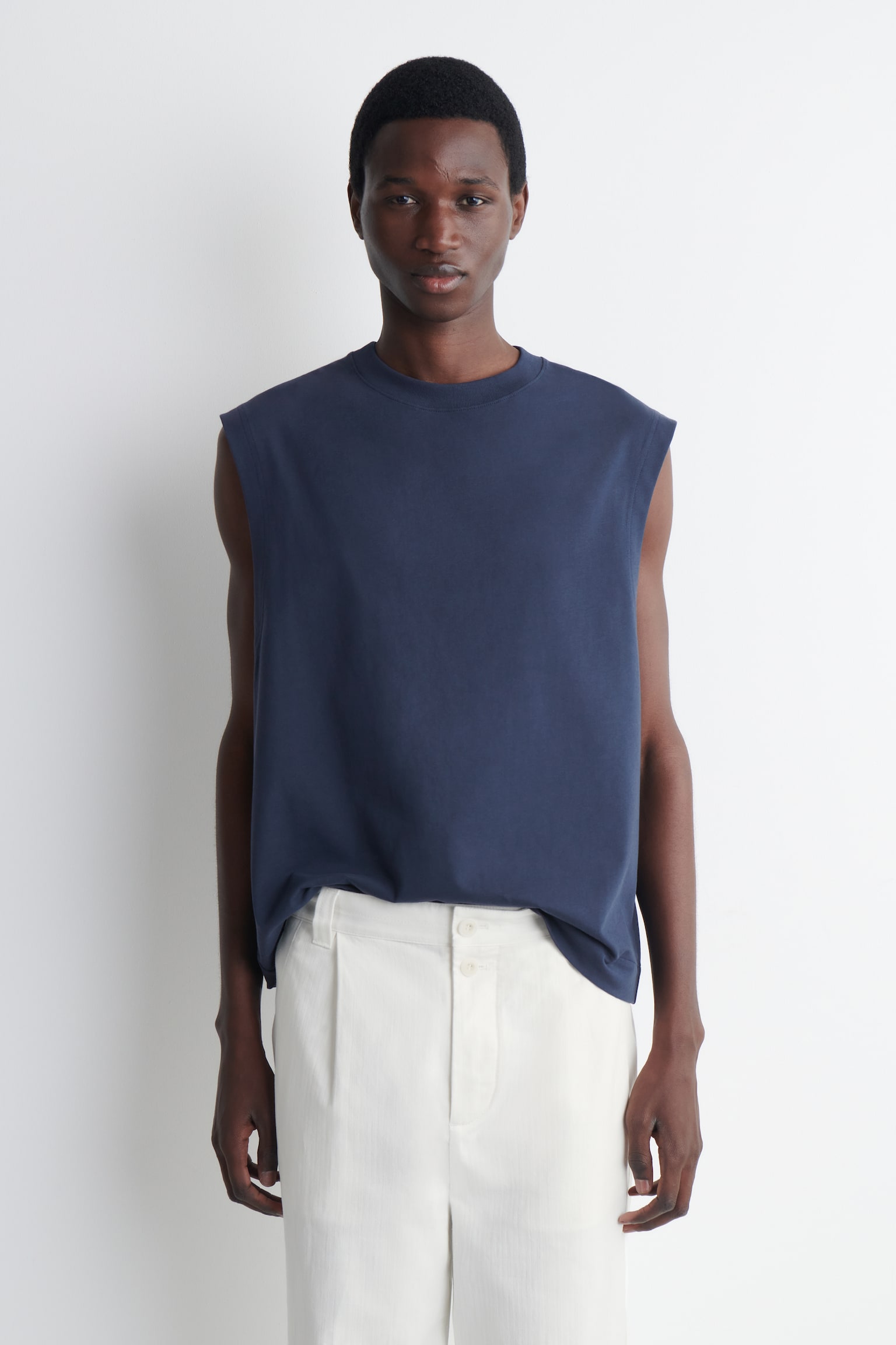 Un haut sans manches bleu marine avec un col ras du cou et une ligne d'épaule étendue, offrant une coupe décontractée lorsqu'il tombe non rentré sur un pantalon clair, face à l'avant. - 1