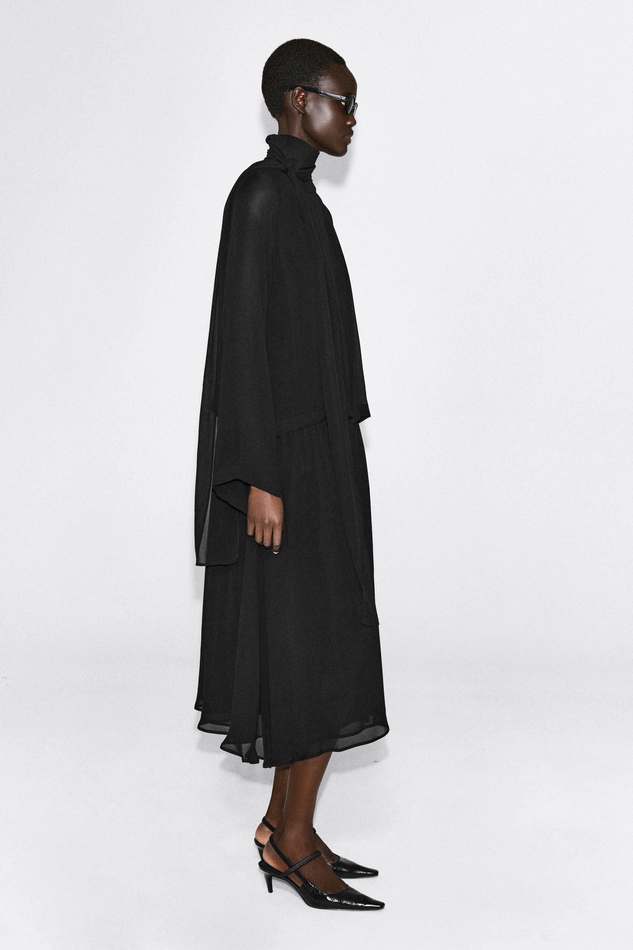 The black fluid scarf-detail midi dress is shown on a model facing right, highlighting its draped, semi-sheer silhouette. The dress features a high wrapped neckline with an integrated scarf detail, long wide sleeves, and a layered, flowing midi-length hem.