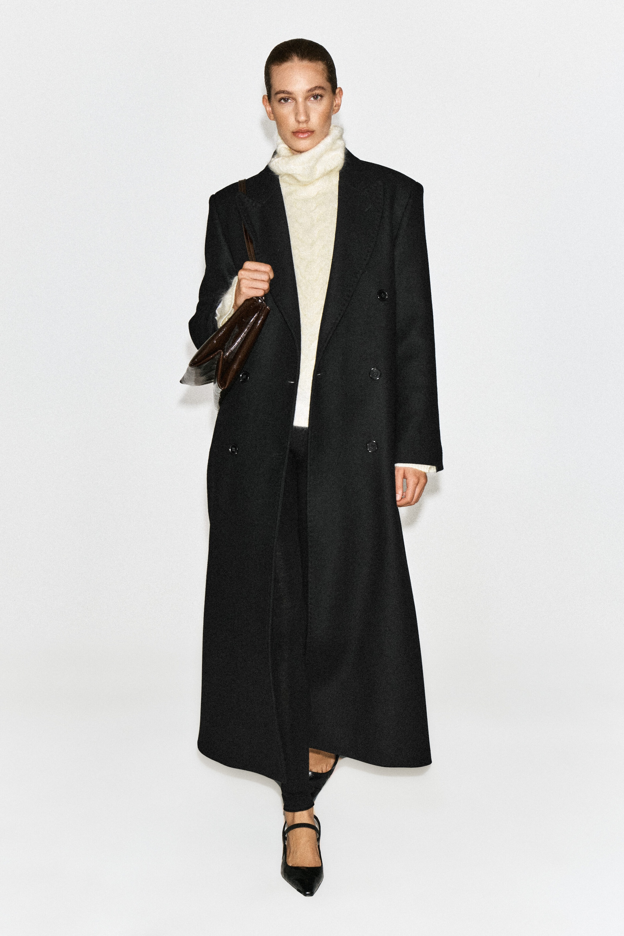 A cream cable-knit brushed cashmere funnel-neck sweater is worn by the model as a layering piece under a long, double-breasted black coat. The sweater, featuring a soft textured knit and a high funnel neck, is styled with black trousers, black pointed-toe heels, and a brown leather shoulder bag.