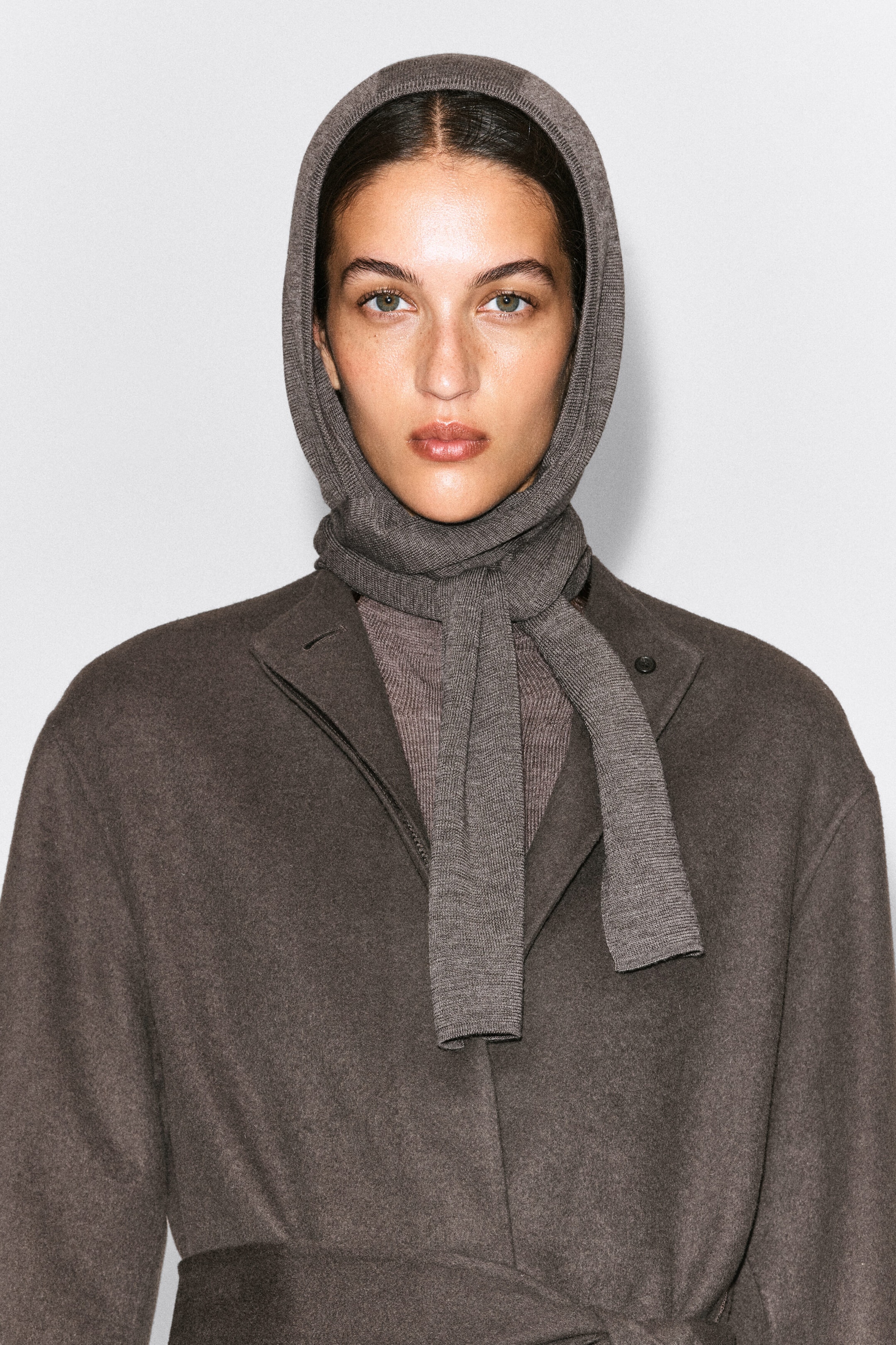 Ein fein gestrickter Kapuzenschal in Taupegrau wird vom Model getragen und bedeckt den Kopf, während er sich um den Hals legt und mit den Enden über die Brust fällt. Das Accessoire ist zusammen mit einem passenden braunen Wollmantel und einem Strickoberteil gestylt. Ein fein gestrickter Kapuzenschal in Taupegrau wird vom Model getragen und bedeckt den Kopf, während er sich um den Hals legt und mit den Enden über die Brust fällt. Das Accessoire ist zusammen mit einem passenden braunen Wollmantel und einem Strickoberteil gestylt.