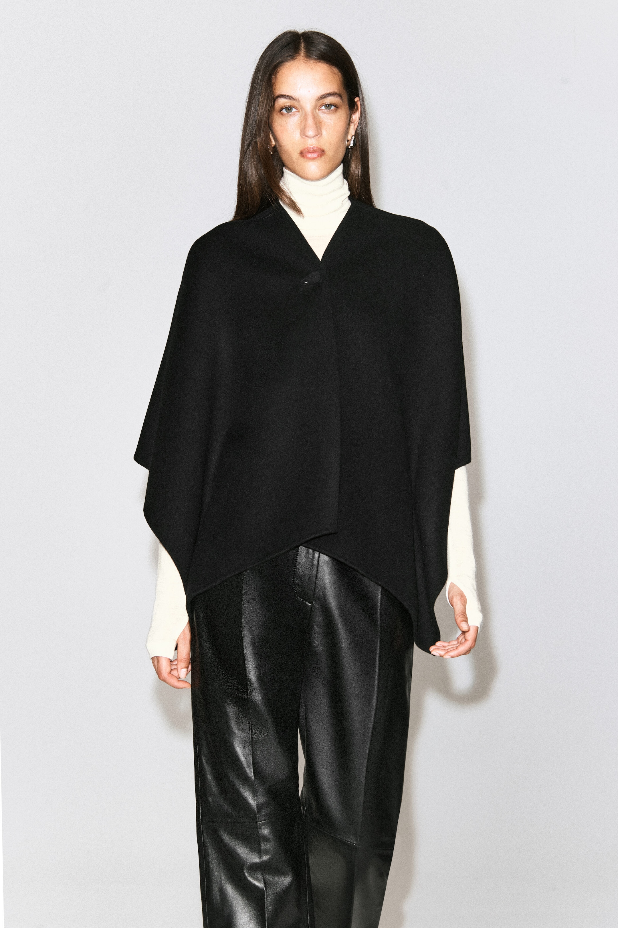 The model is wearing a black double-faced wool-blend cape, featuring a V-neckline with a single top-button fastening and a draped, asymmetric hem. The cape is layered over a cream-coloured roll-neck sweater and paired with black leather trousers. The model is wearing a black double-faced wool-blend cape, featuring a V-neckline with a single top-button fastening and a draped, asymmetric hem. The cape is layered over a cream-coloured roll-neck sweater and paired with black leather trousers.