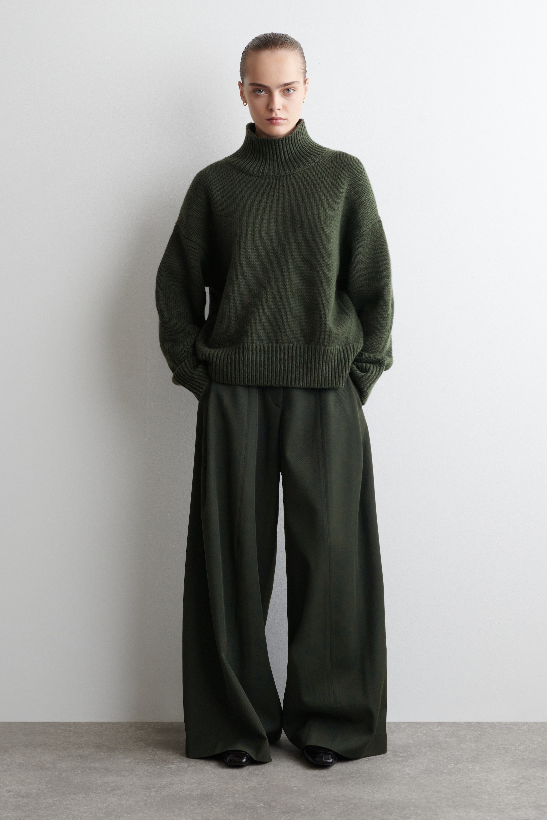 Größeres Bild anzeigen: Ein dunkel olivgrünes monochromatisches Outfit wird gezeigt, bestehend aus einem übergroßen Rollkragenpullover und weiten Hosen. Der Pullover zeichnet sich durch einen gerippten Rollkragen und Saum aus, während die Hosen eine entspannte, fließende Silhouette mit einer sichtbaren zentralen Naht entlang jedes Beins aufweisen.